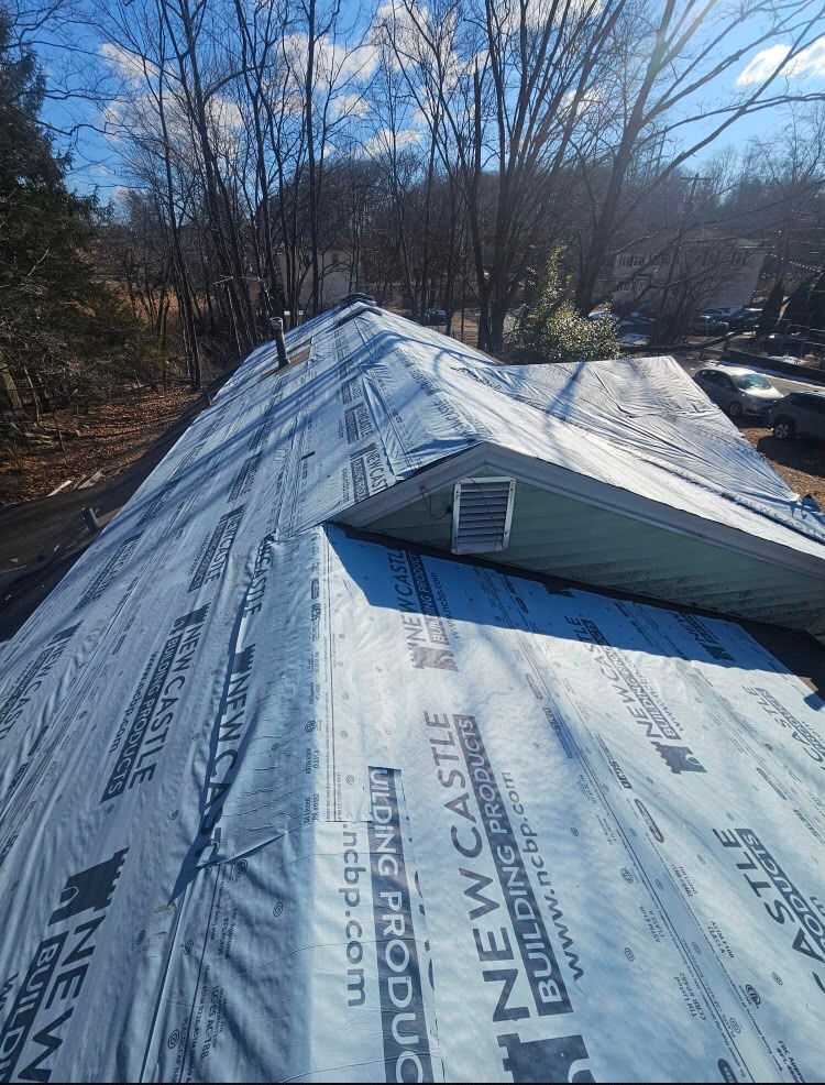Rooftop under renovation with blue New Castle underlayment and a small vent in a wooded residential area