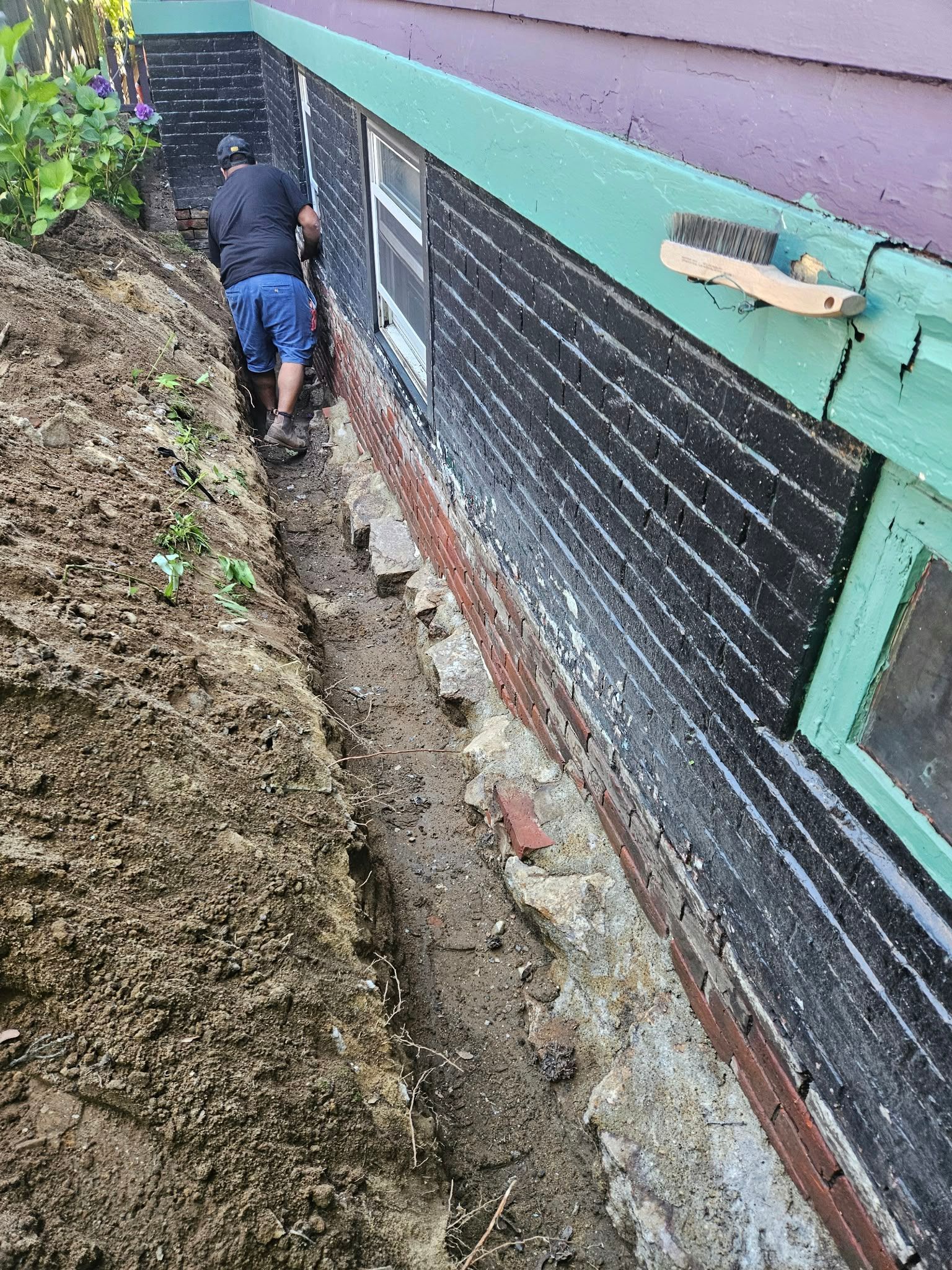 Worker digging a narrow trench beside a house with black siding and green trim