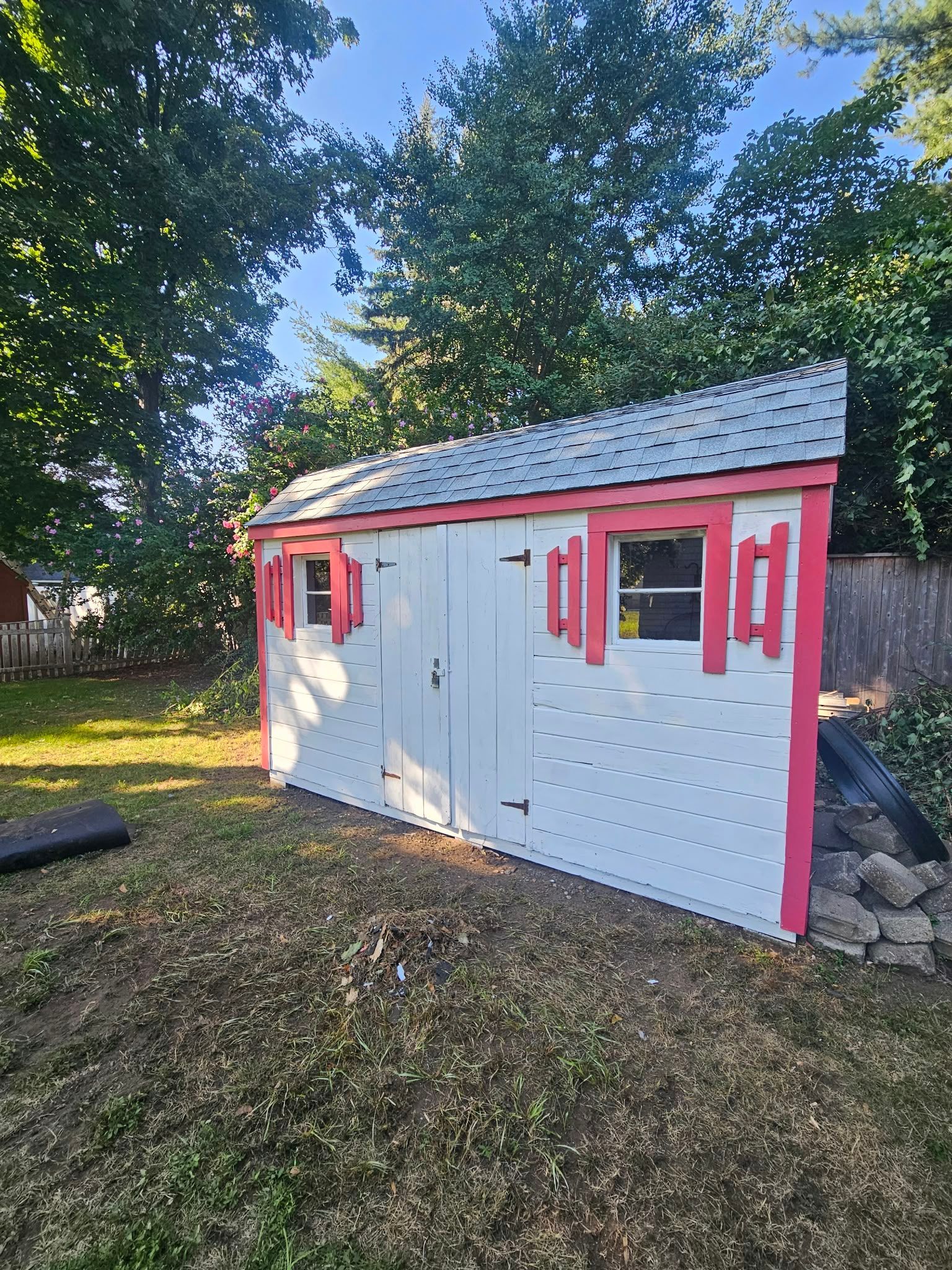 Small white shed with pink trim and windows in a leafy backyard, beside a stone wall
