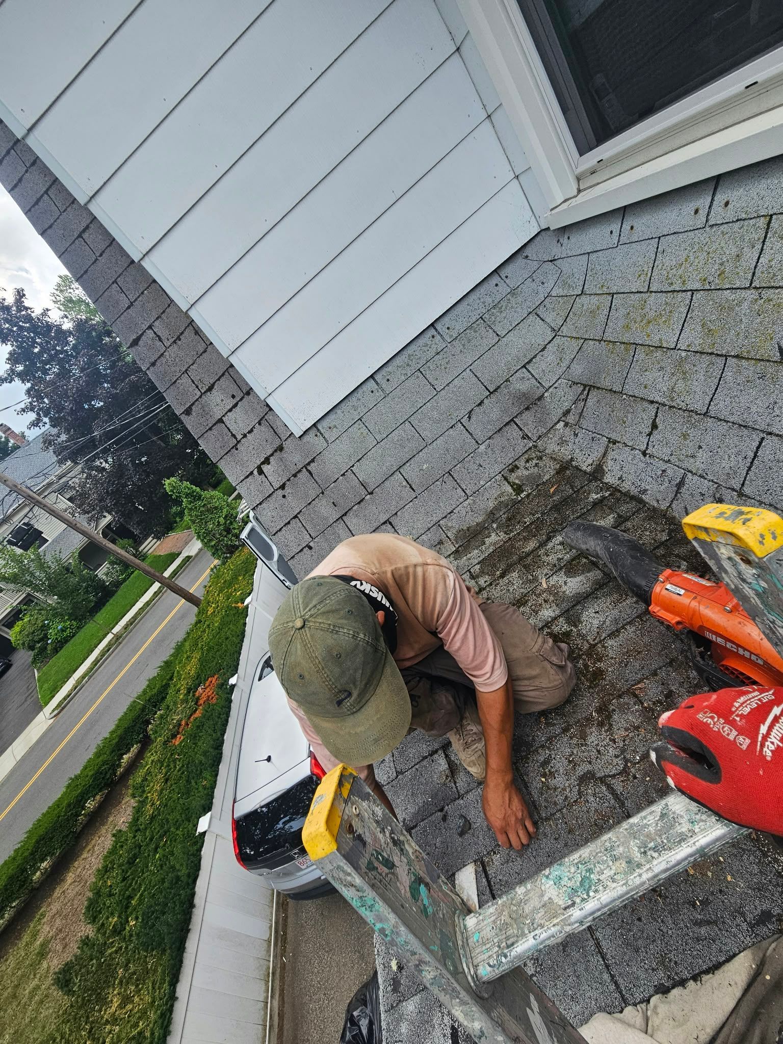 Worker on a roof cleaning shingles with tools near a white house and red ladder.