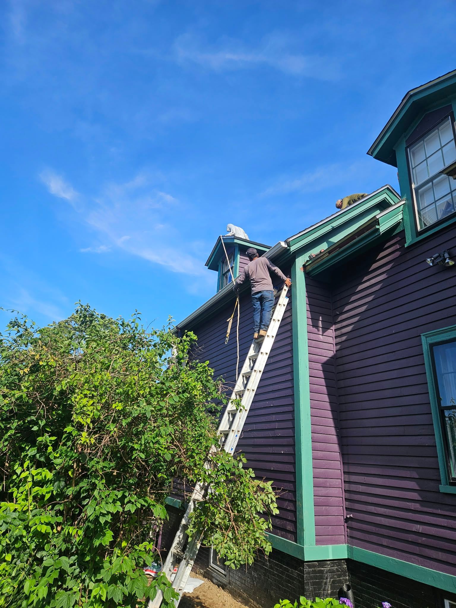 Worker on a ladder repairing a dark purple house roof under a bright blue sky