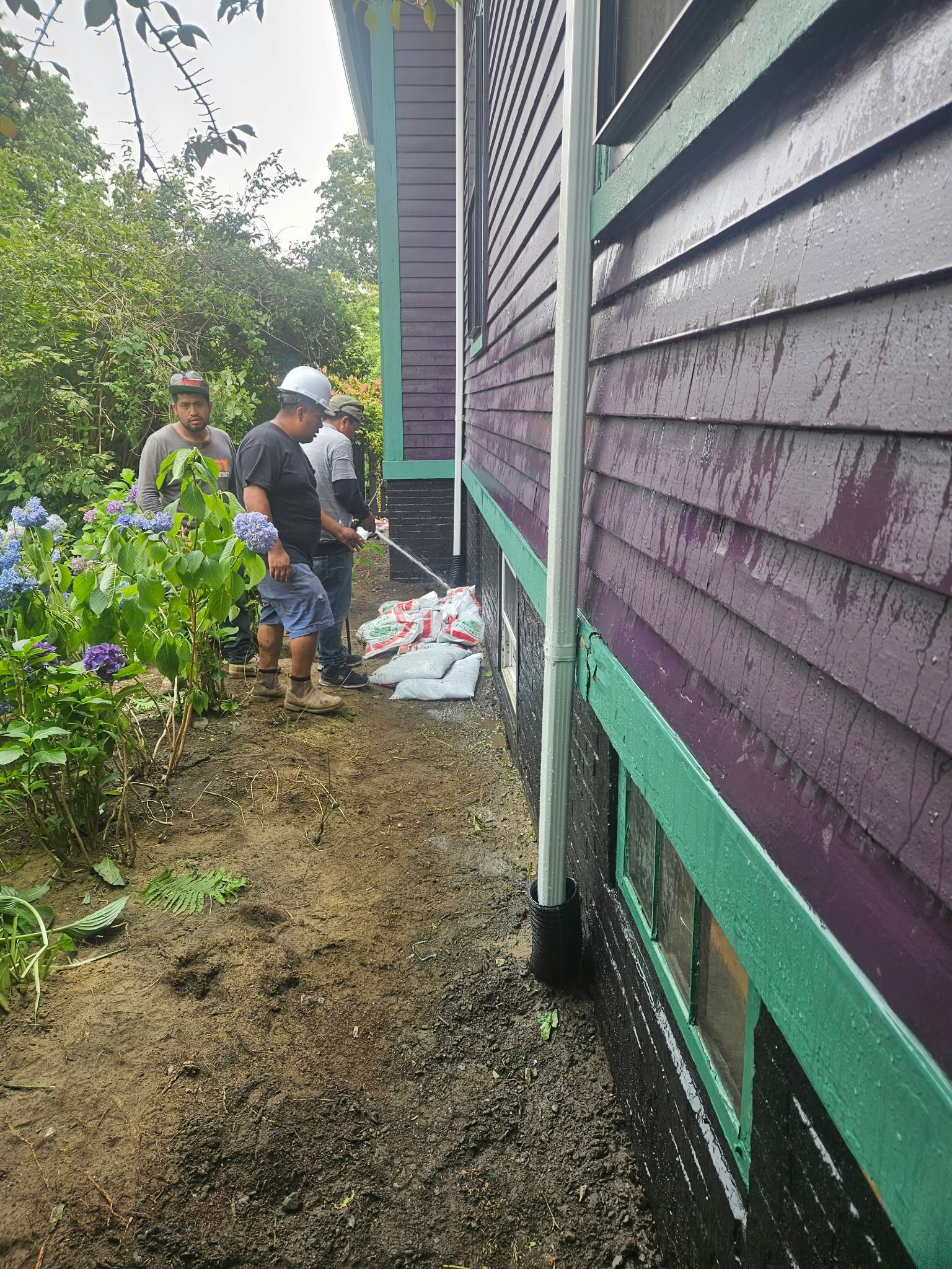 Two workers by a purple house clearing debris along a muddy side path
