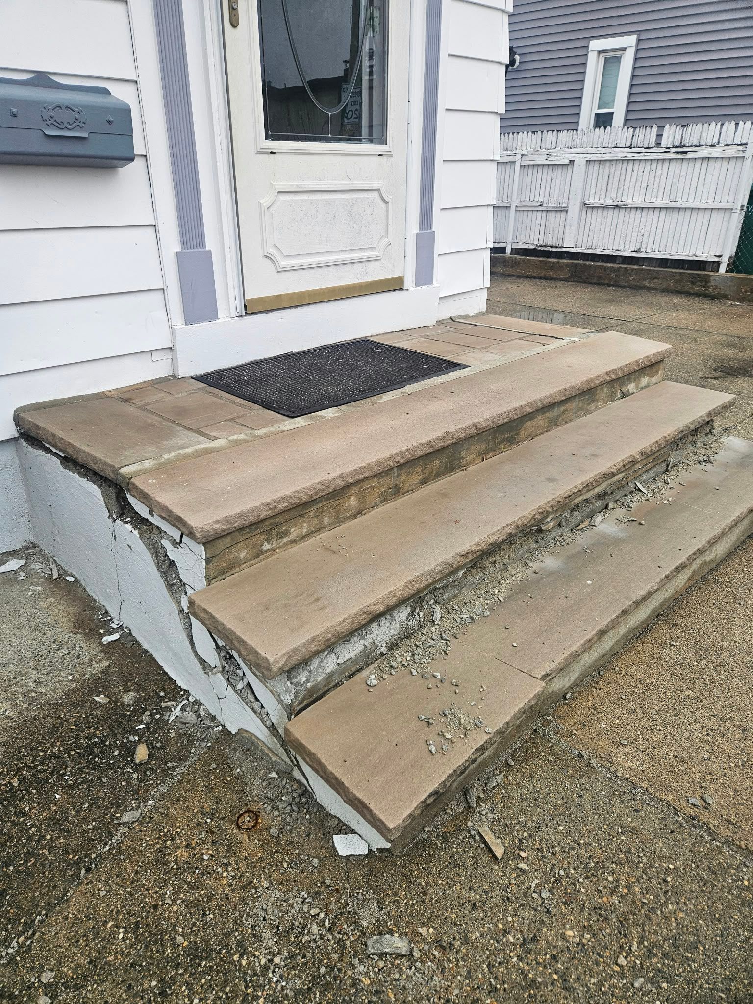 Concrete front steps with a white door and black doormat, showing chipped and weathered edges
