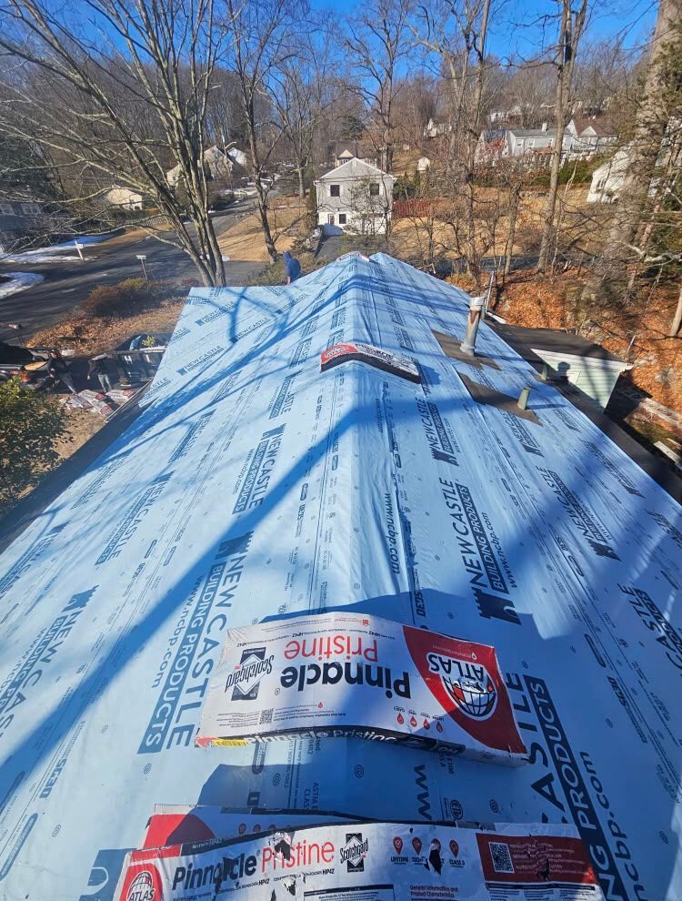 Blue-roofed house under construction with roofing underlayment and a wooded neighborhood view