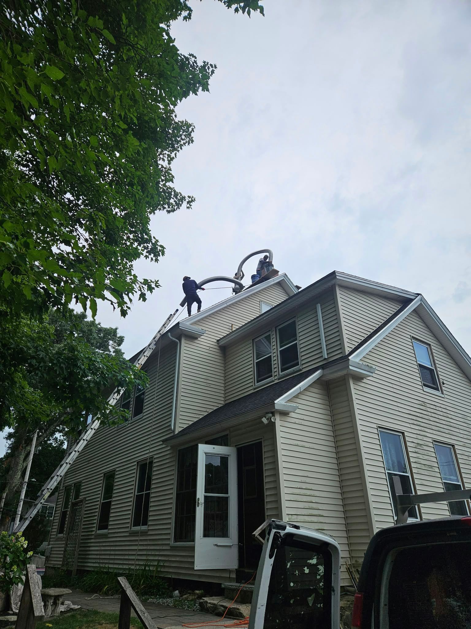 Damaged house with workers on the roof beside a large tree, viewed from the driveway