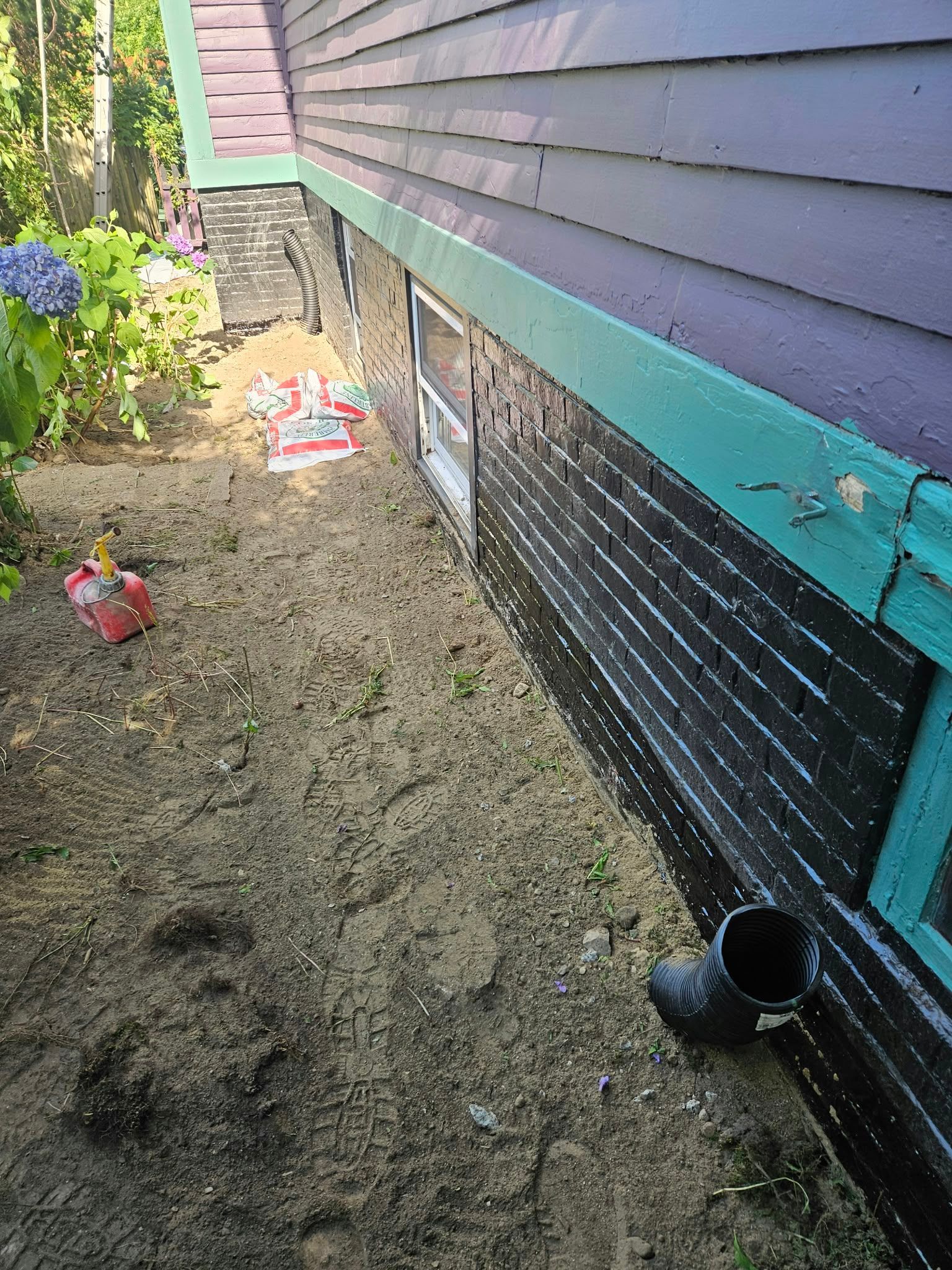 Narrow dirt trench along a house foundation with exposed black waterproofing and teal trim.