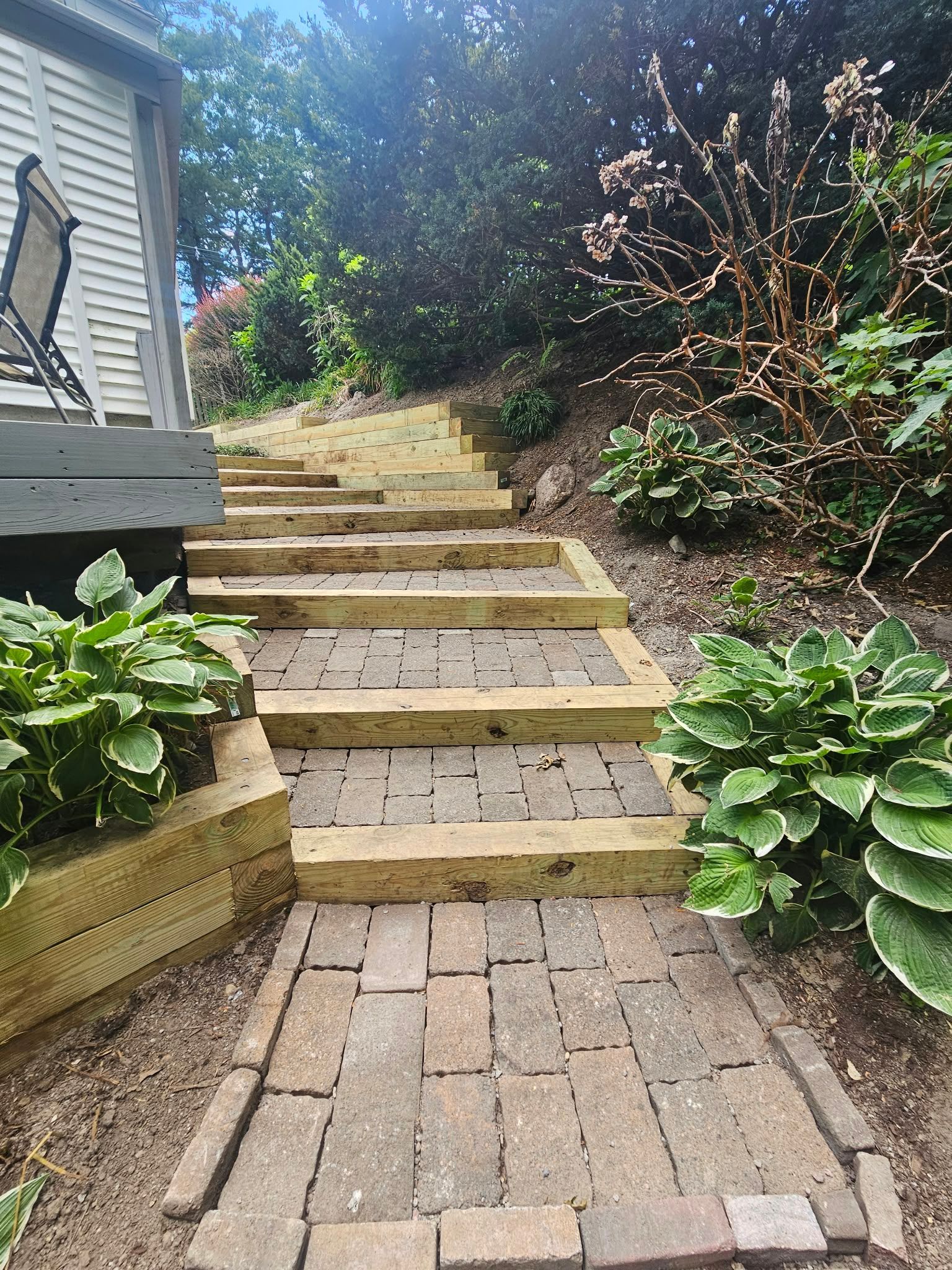 Brick path leading up a steep stone stairway beside a house, with green plants and flowering shrubs.