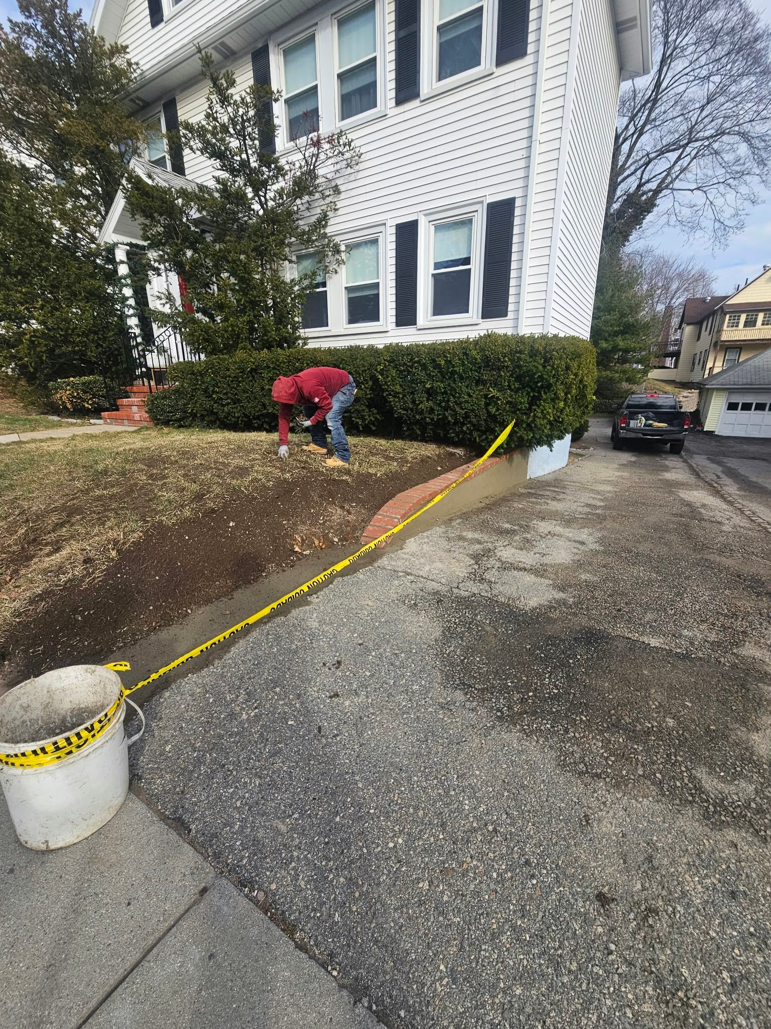 Person in red shirt weeding a sloped garden beside a white house and driveway.