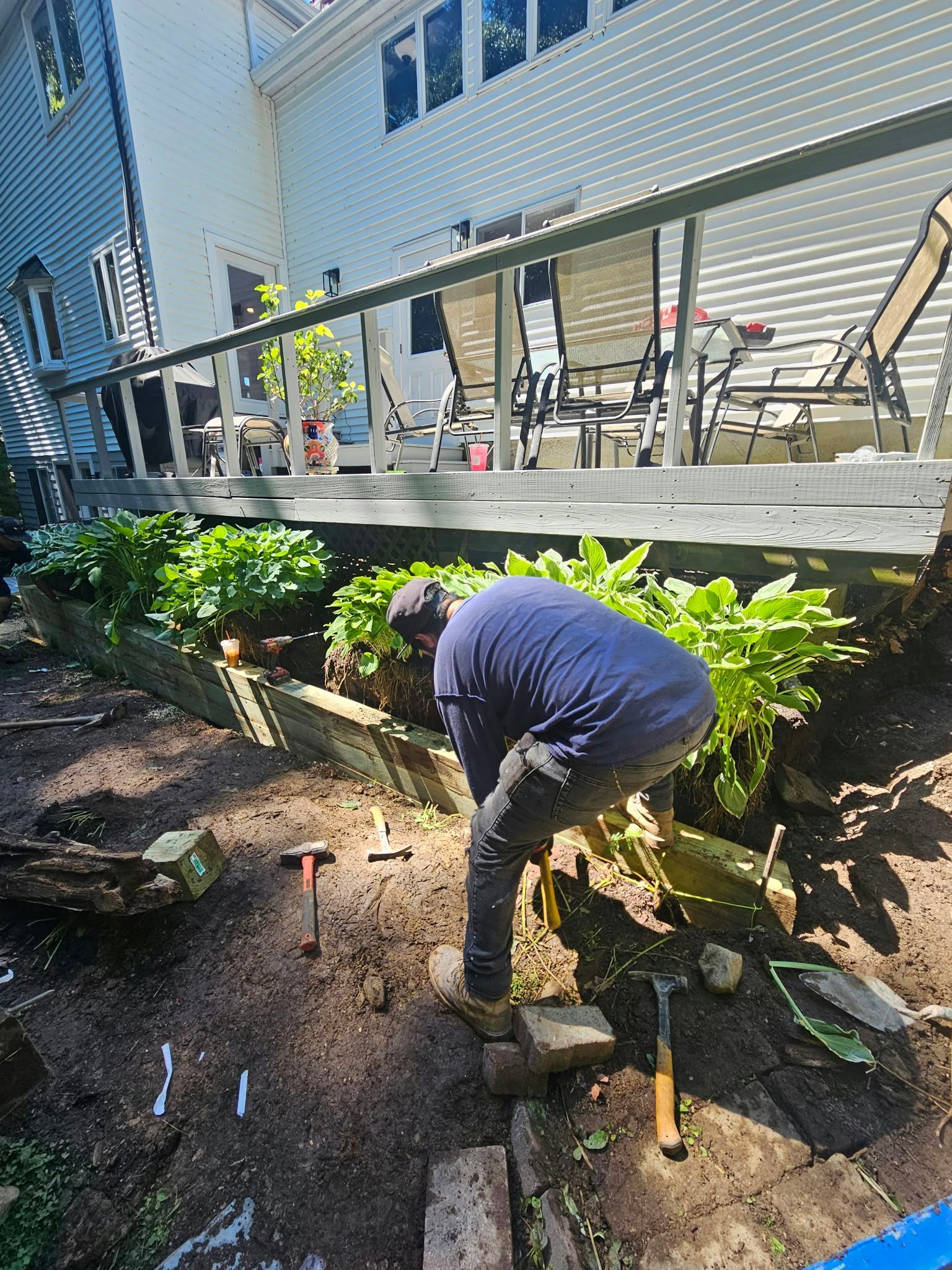 Worker digging beside a building patio, with tools and green shrubs in the foreground.