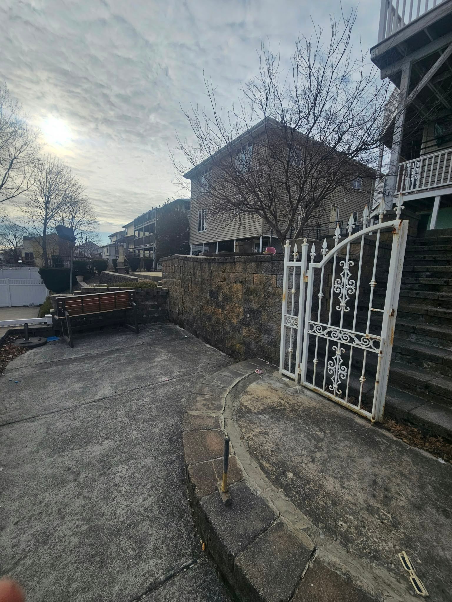Quiet residential street with stone wall, white gate, parked car, and houses under a cloudy sky