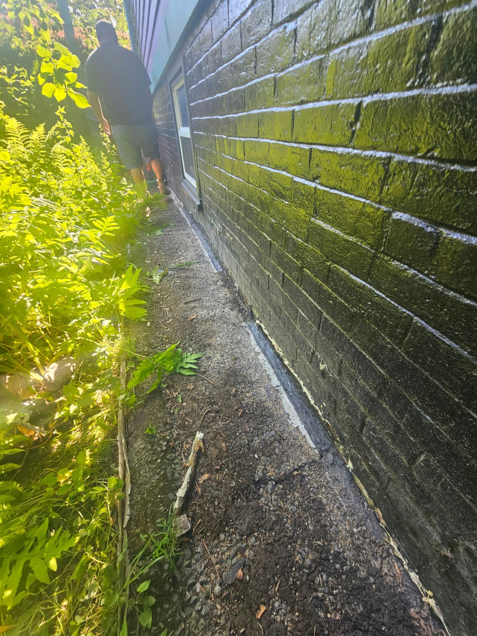 Person walking beside a sunlit brick wall along a narrow gravel path with green foliage nearby