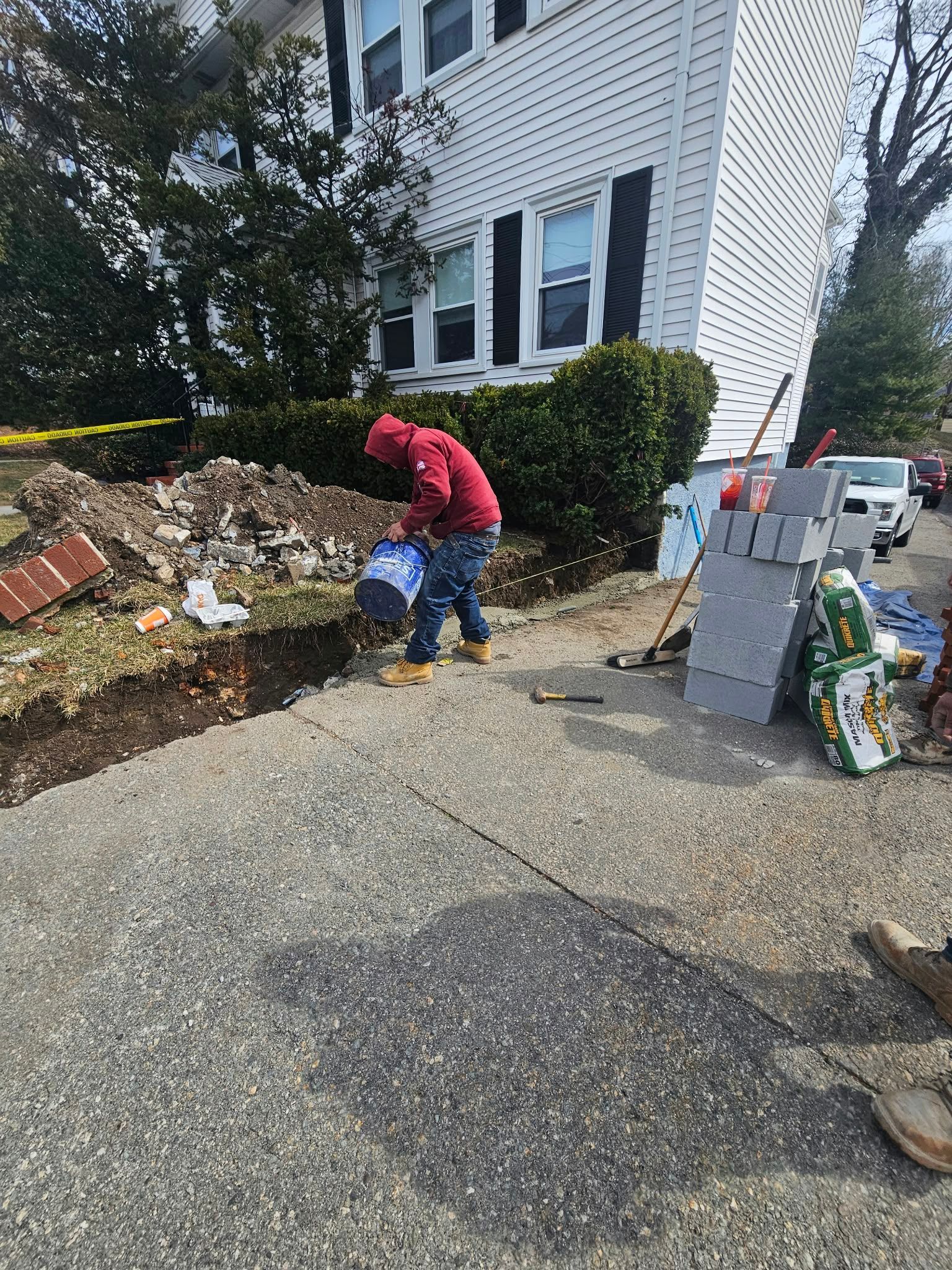 Person in red jacket bending near a blue bin by a house driveway with debris and tools nearby