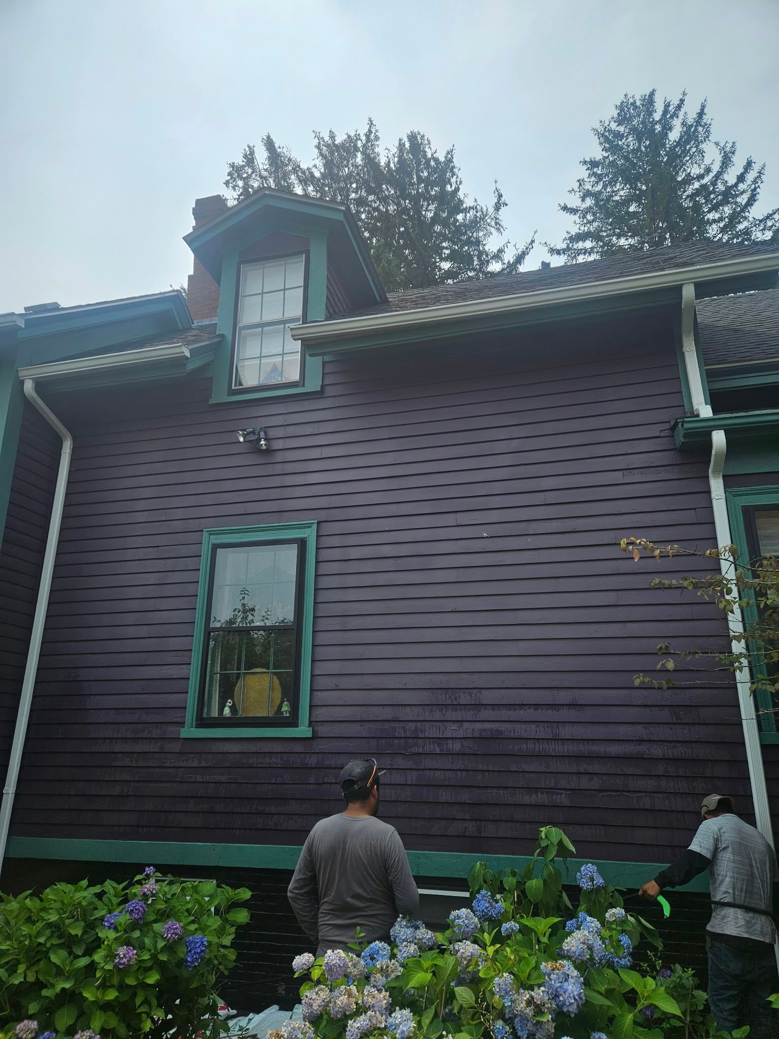 People standing beside a purple house with green-trimmed windows and flowers in the foreground.