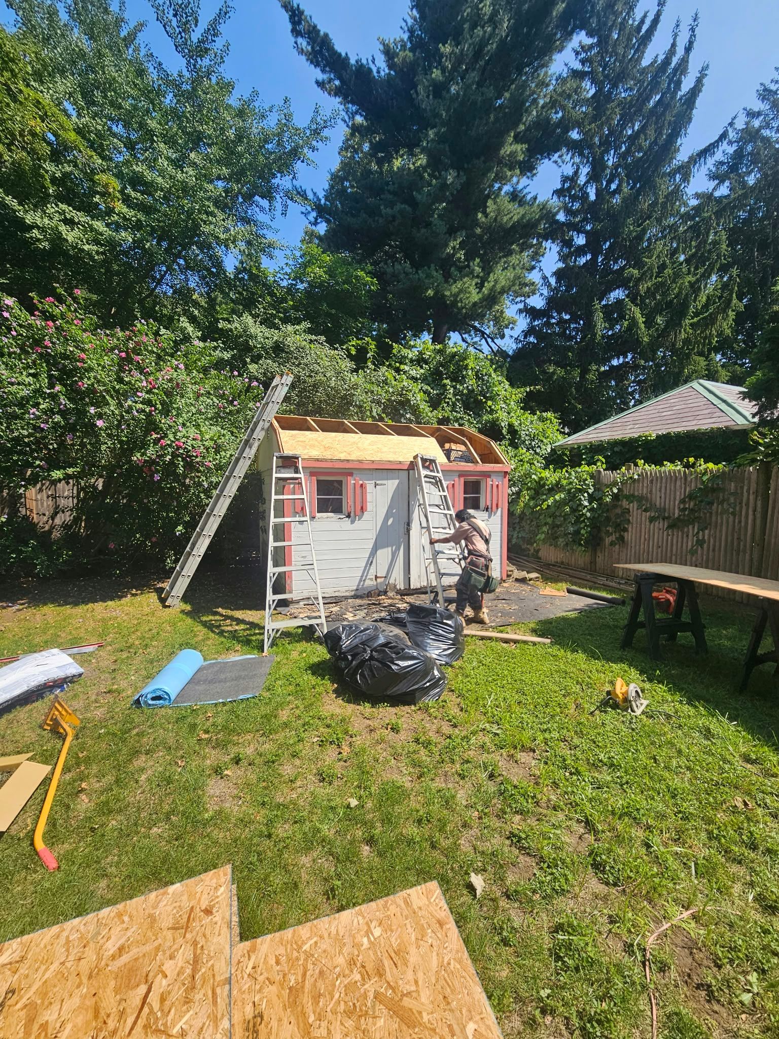 Backyard playhouse under tall trees, with lawn tools and a table nearby.