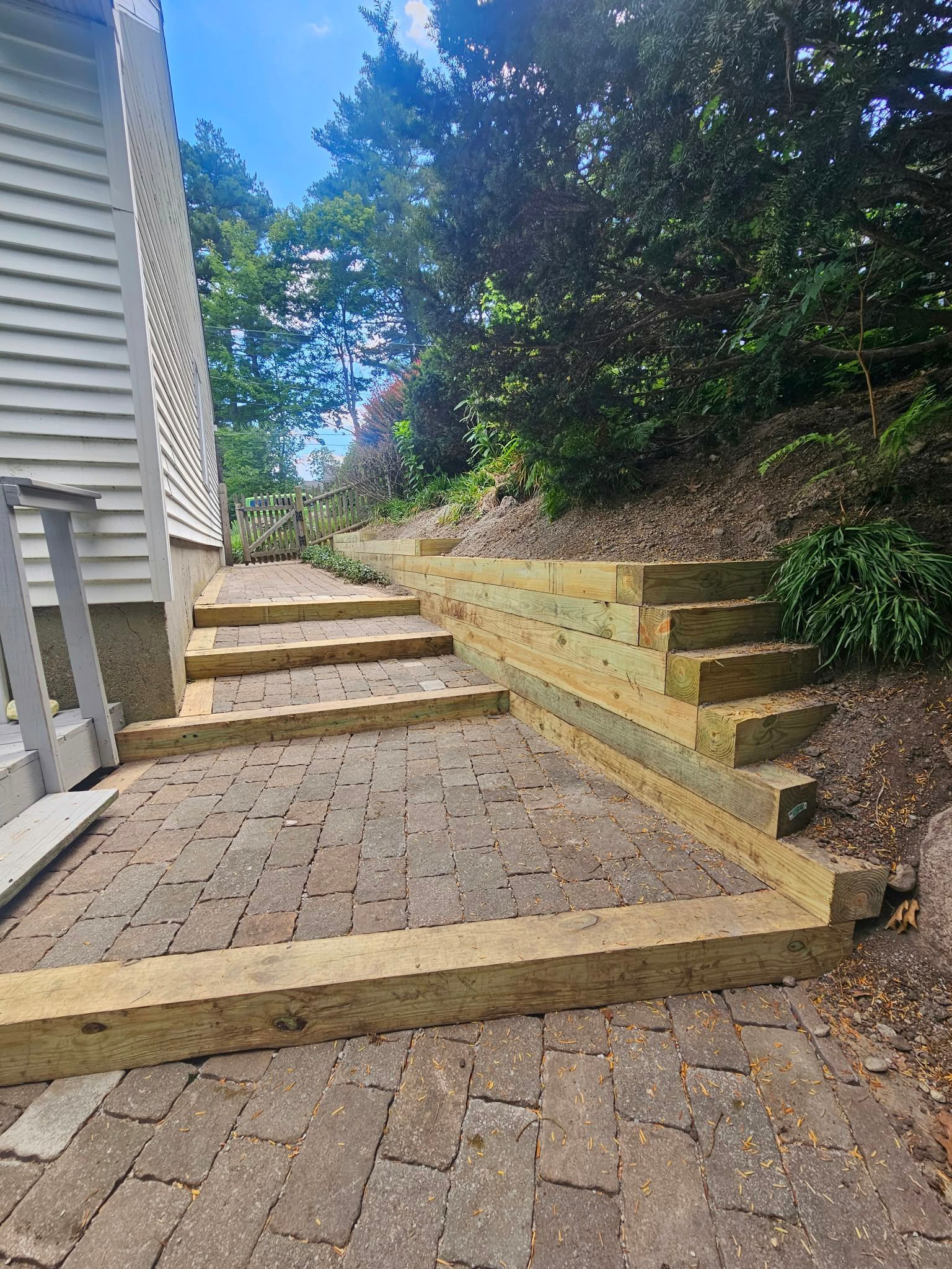 Brick walkway with yellow-edged steps beside a house and retaining wall on a sunny hillside