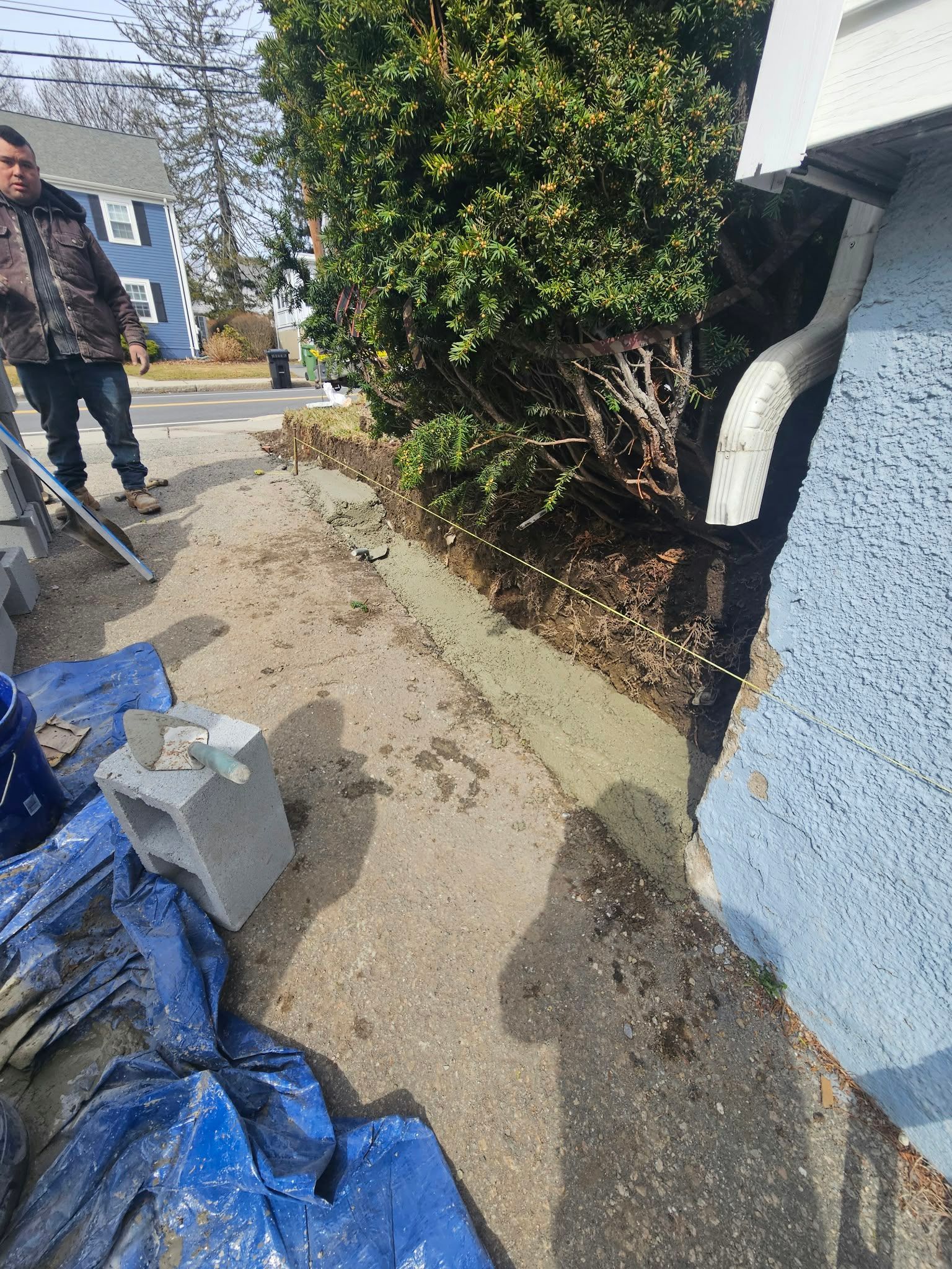 Driveway with blue tarp, debris, and a person using a shovel beside a house and hedge.