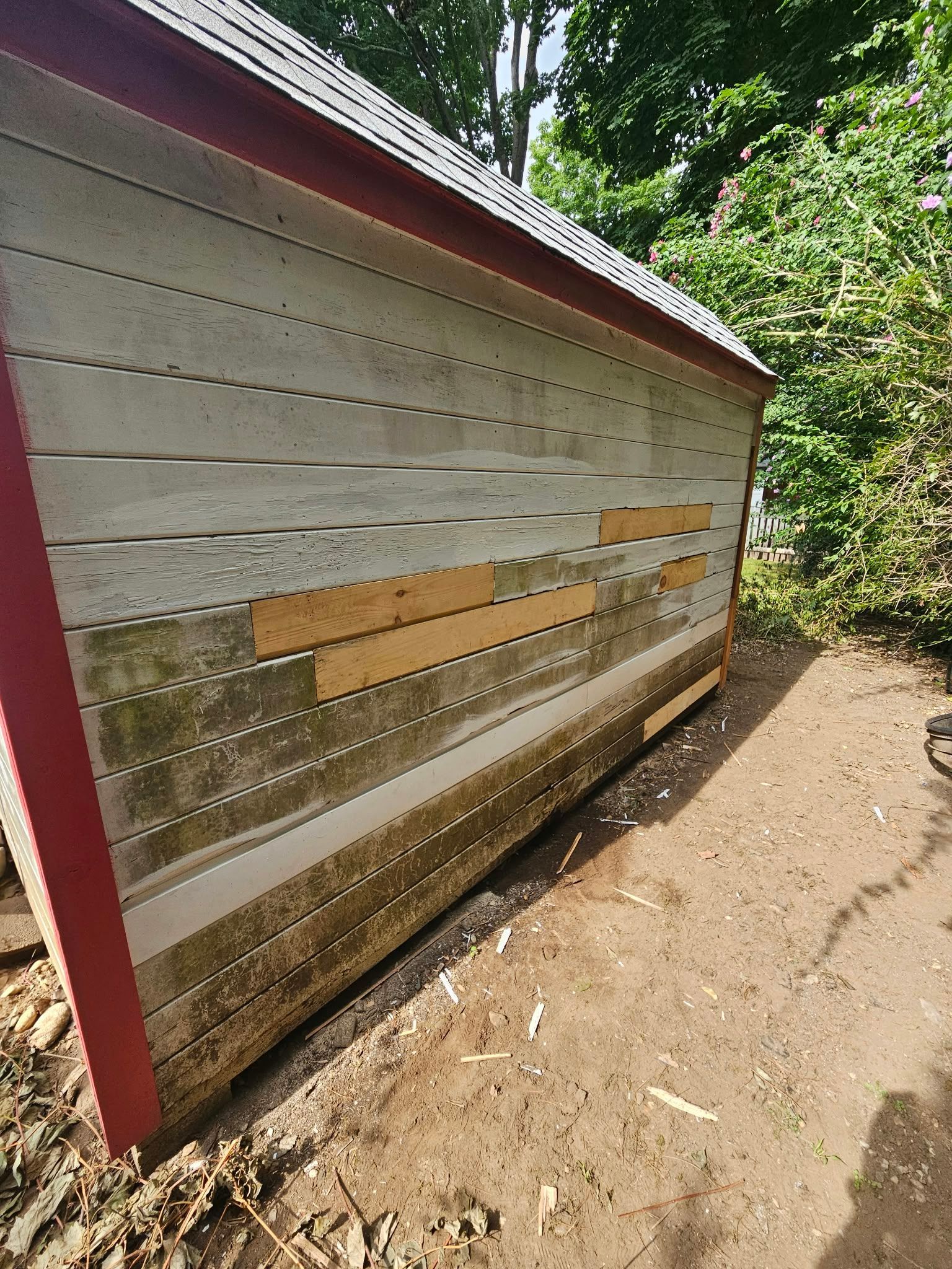 Damaged red-trimmed exterior wall with missing and broken siding beside a dirt path.