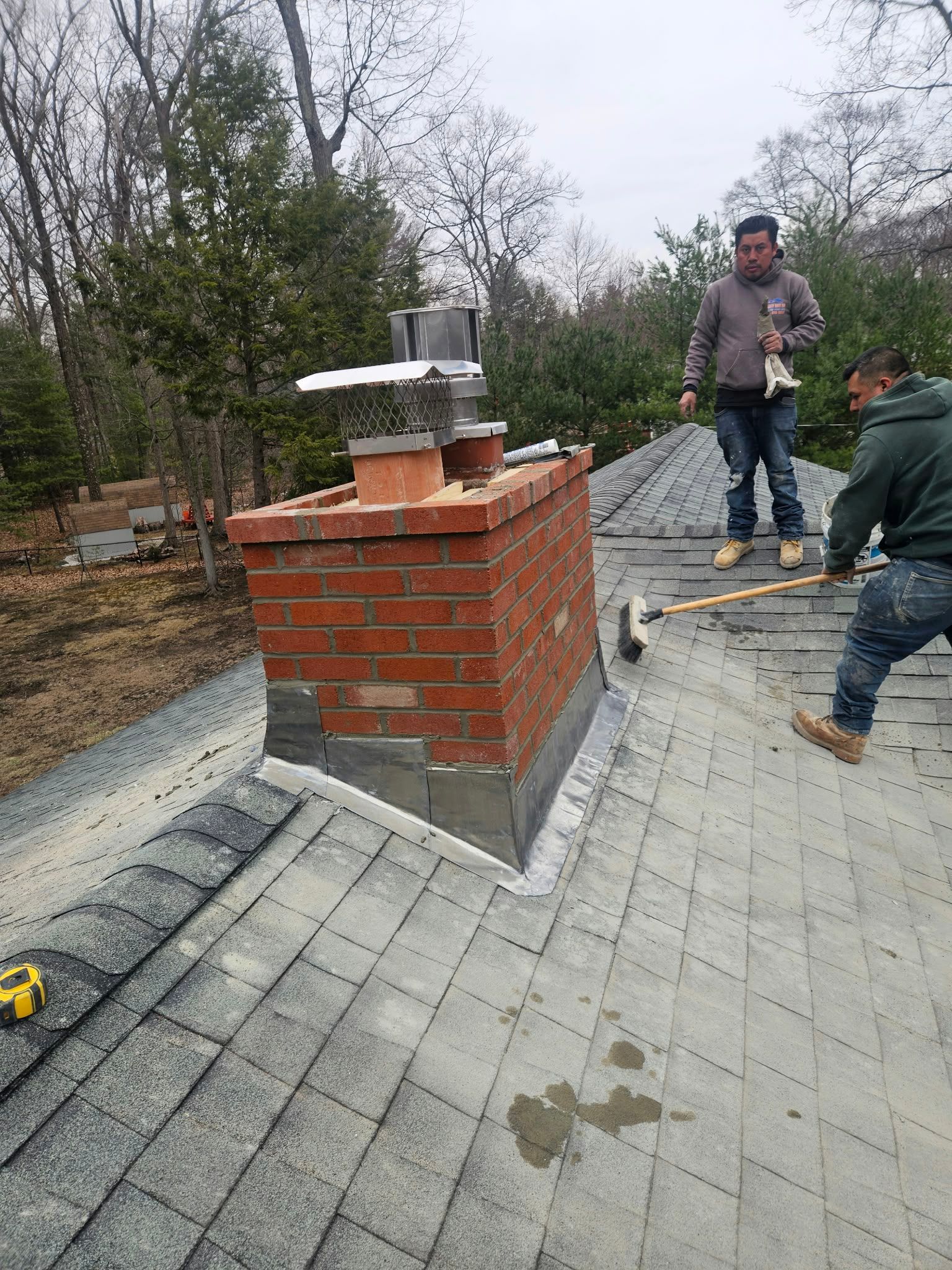 Workers inspecting a brick chimney on a rooftop under construction, with tools and a sloped roof nearby.