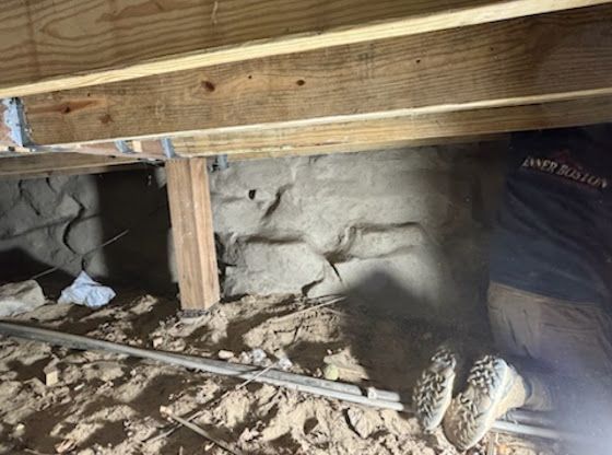 Crawlspace under a house with exposed beams, stone foundation, debris, and a worker’s boot visible on the right