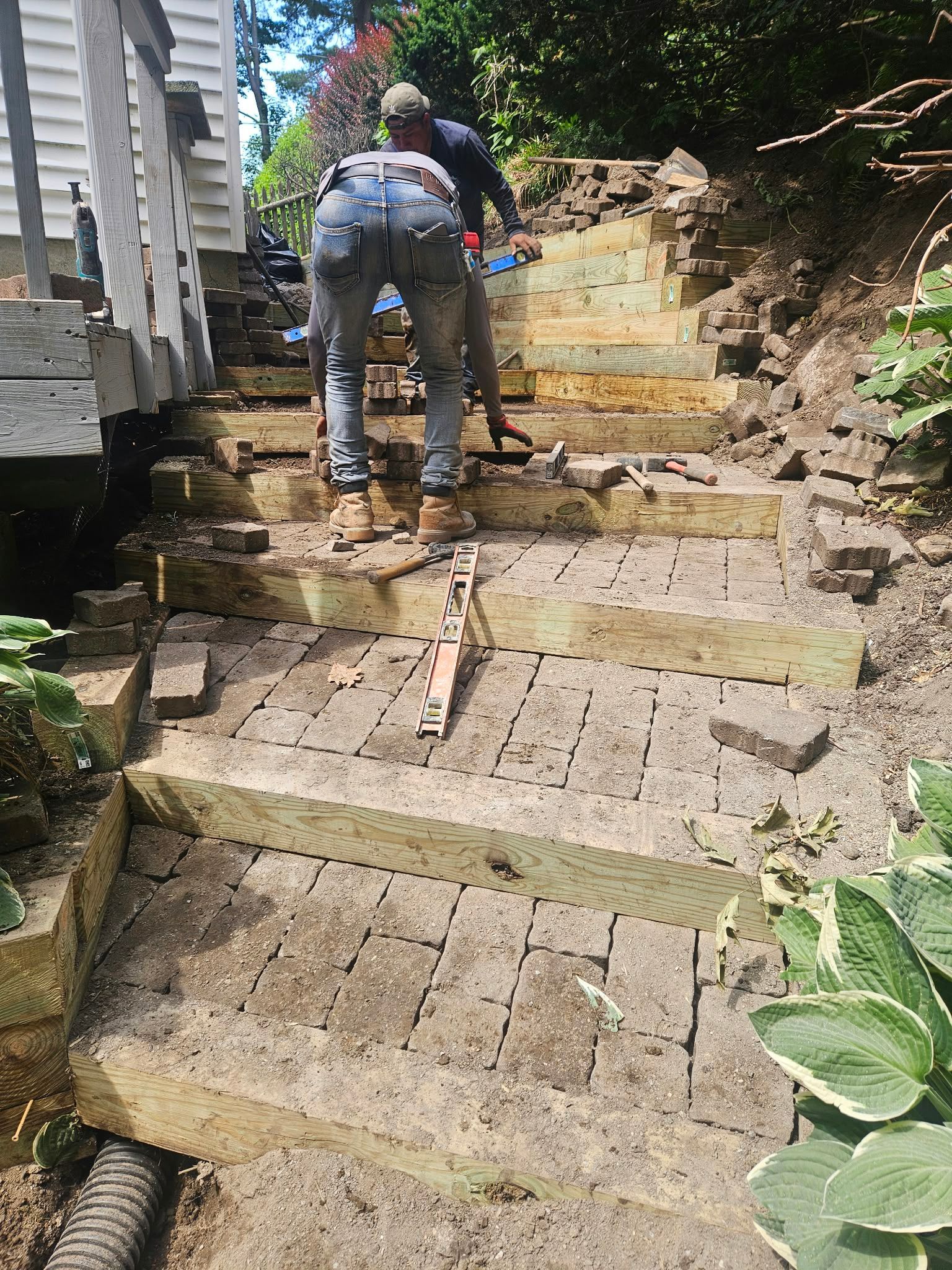 Two workers repairing stone steps beside a house and dirt hillside.