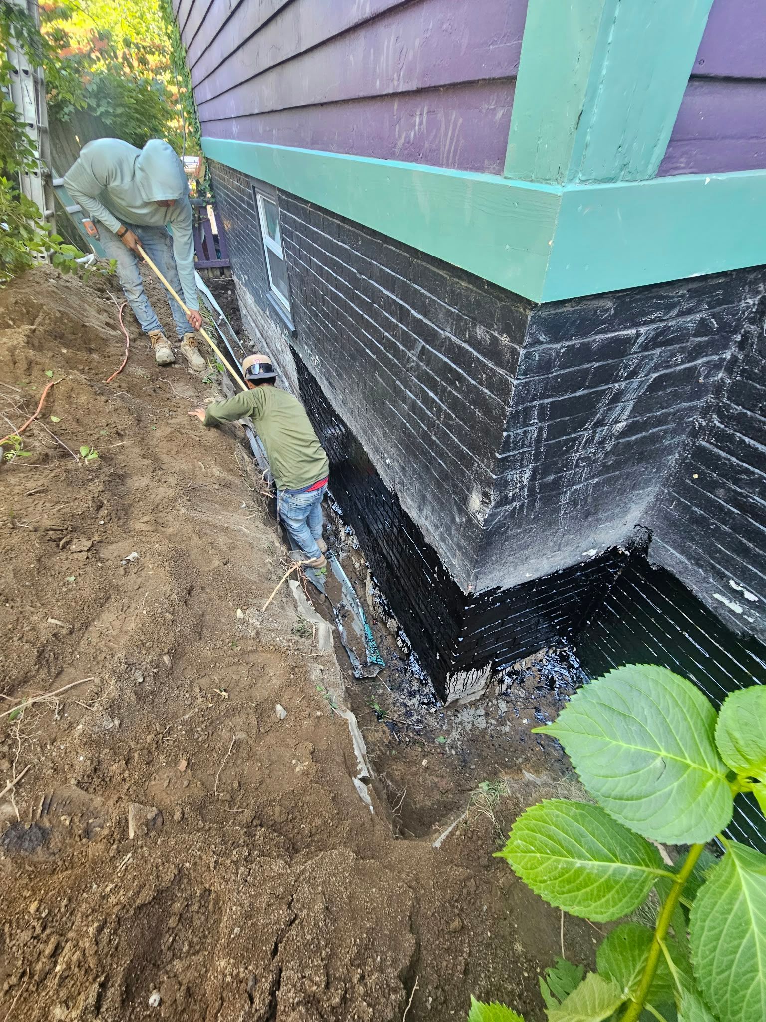 Workers digging a trench along a house foundation with black waterproofing and green siding.