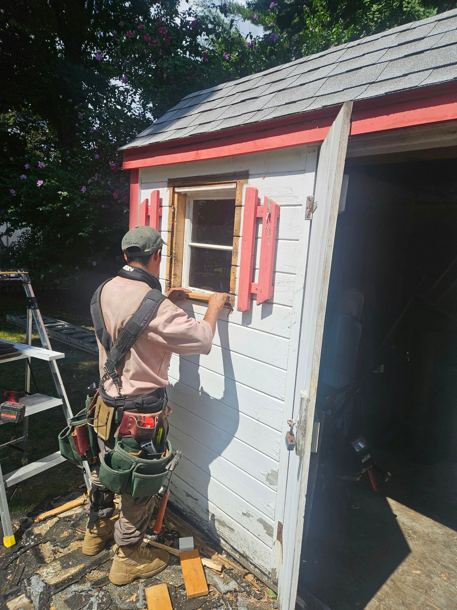 Worker painting trim on a small gray shed with red accents outdoors.