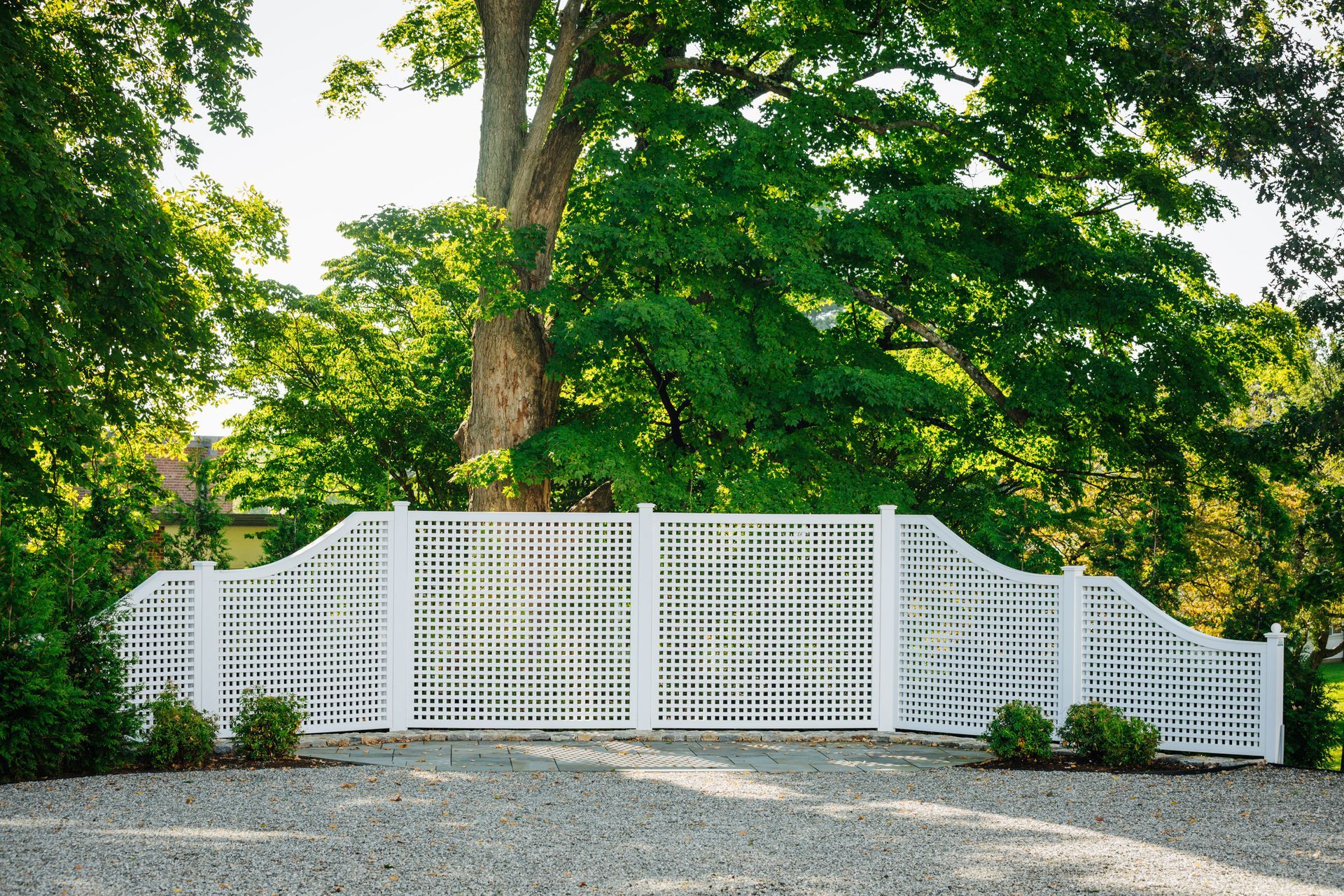 A white fence is surrounded by trees in a gravel driveway.