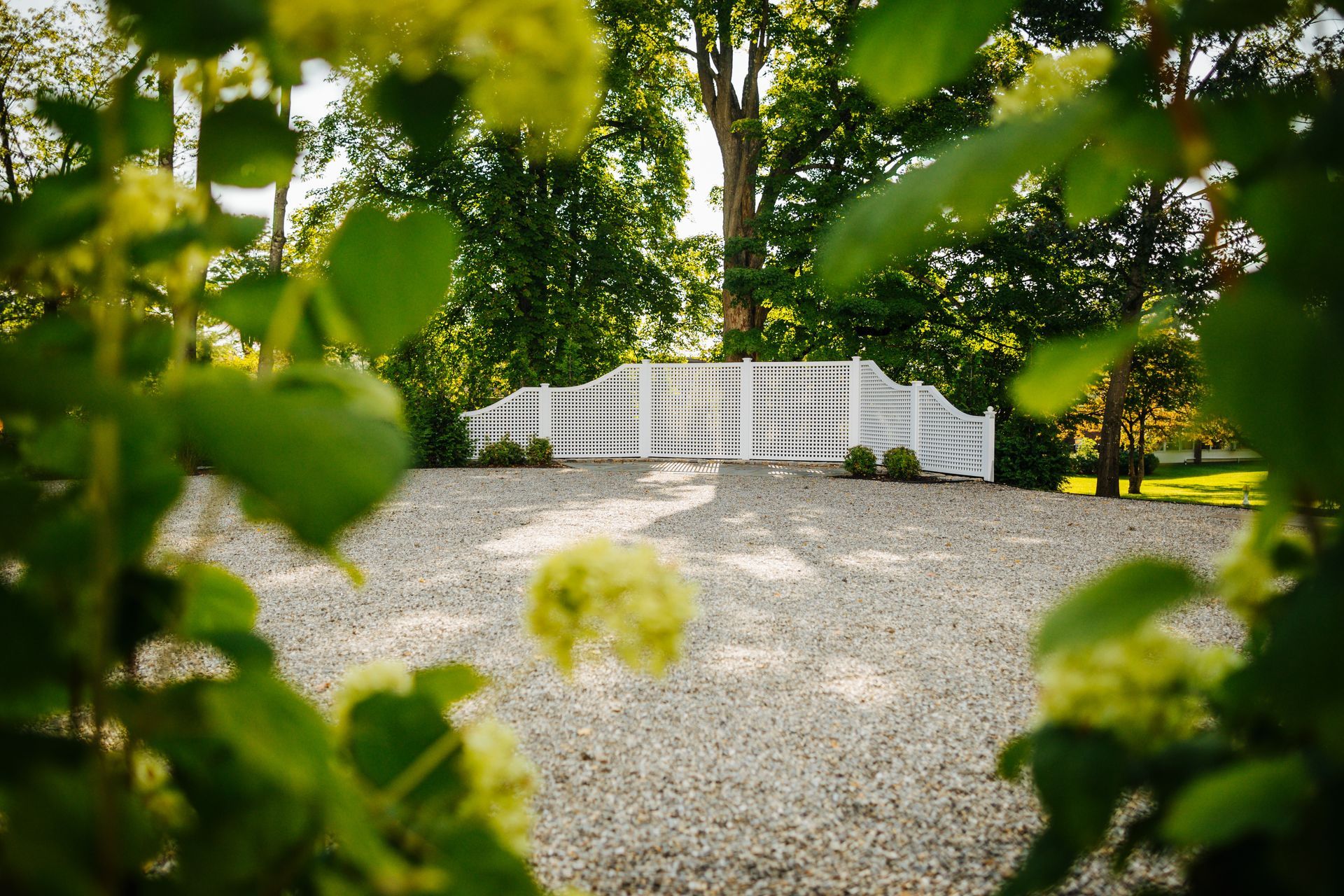 A white fence is surrounded by trees and flowers in a gravel driveway.