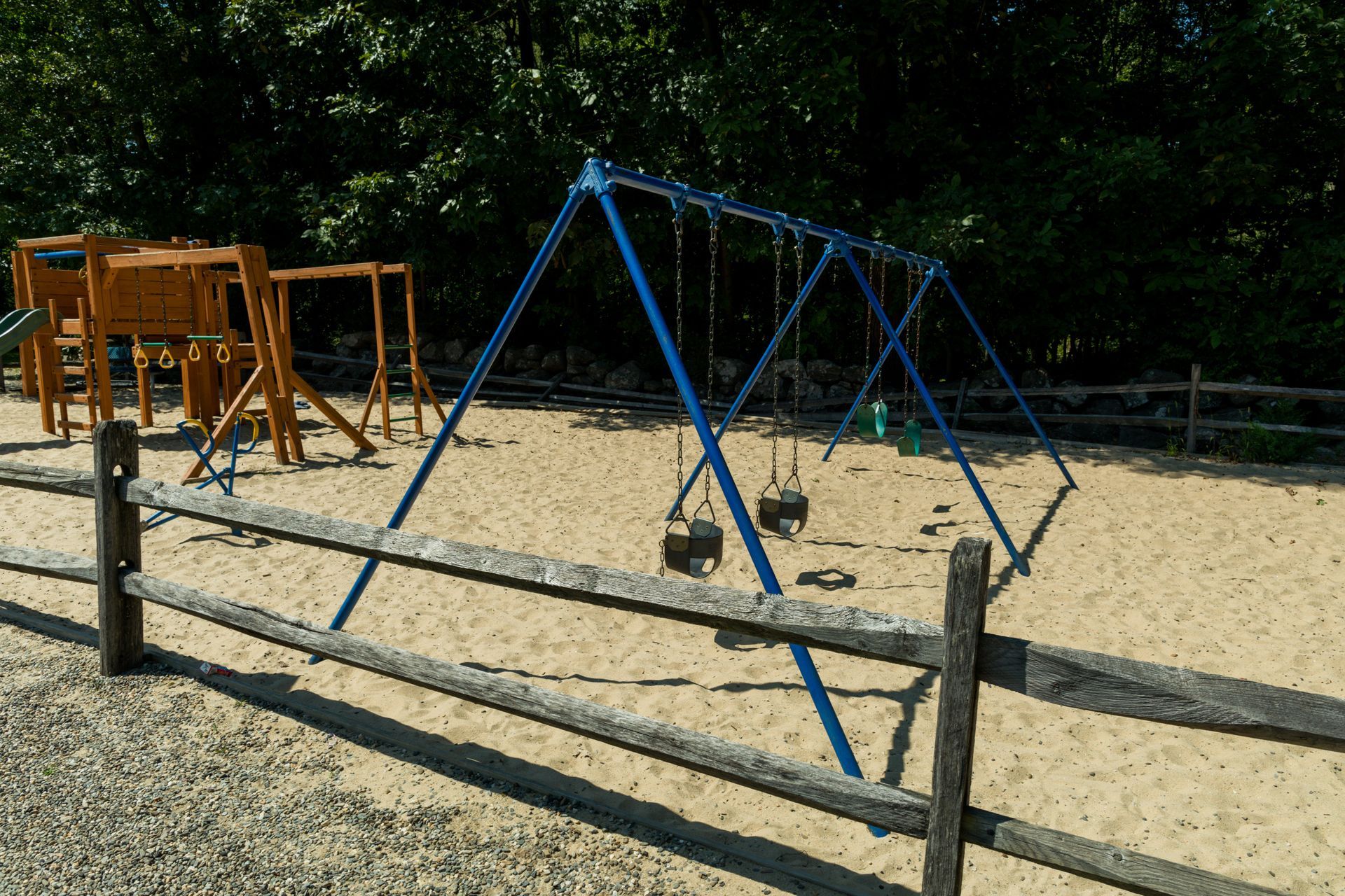 A wooden fence surrounds a playground with swings in the sand.