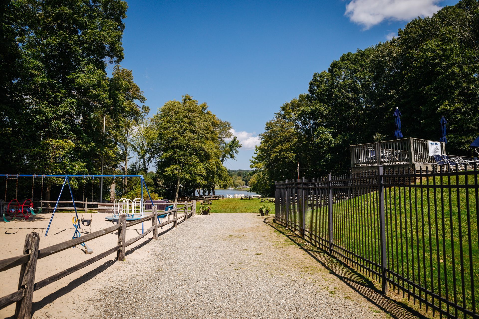 A gravel road leading to a playground surrounded by trees and a fence