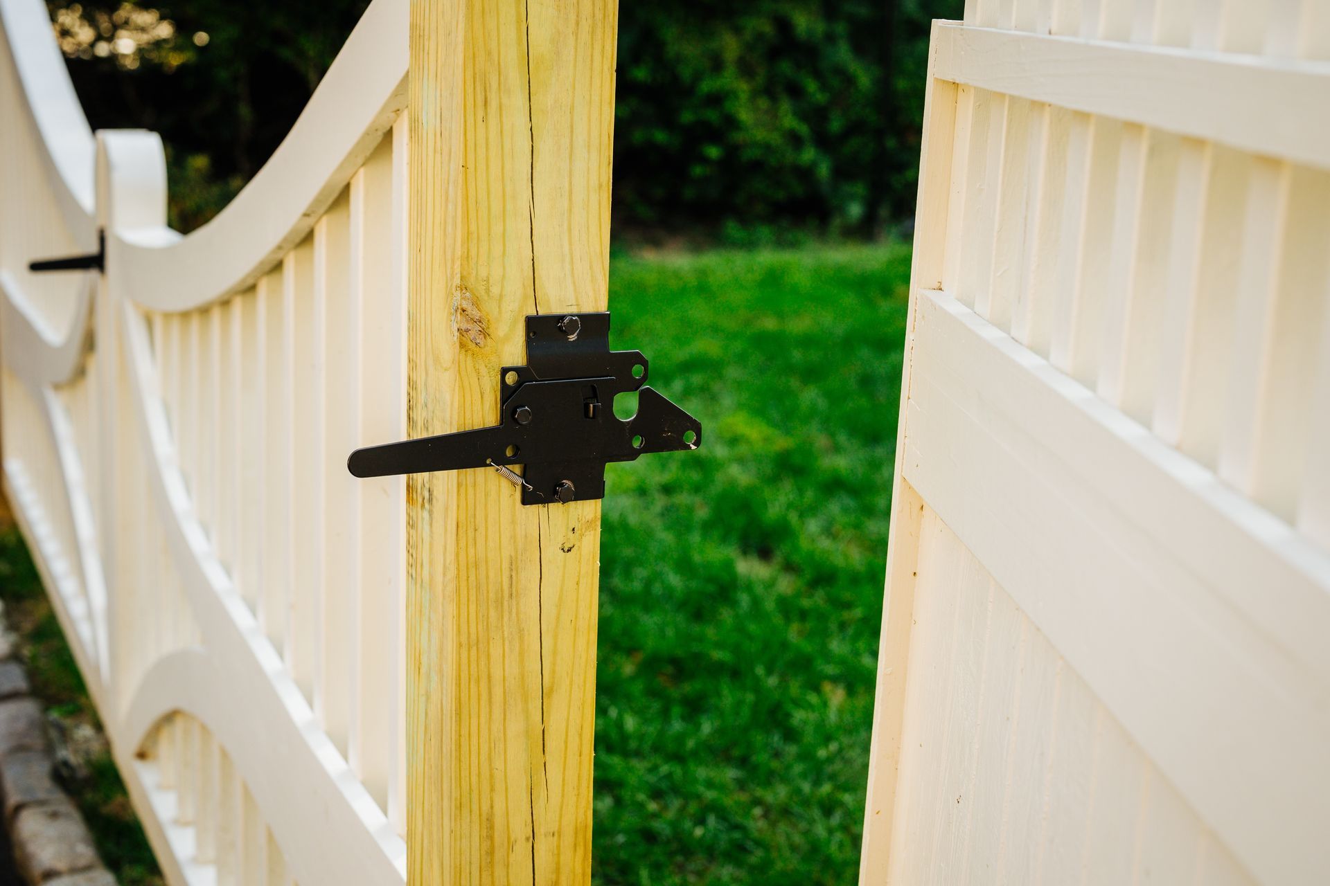 A white fence with a black latch is open to a grassy yard.