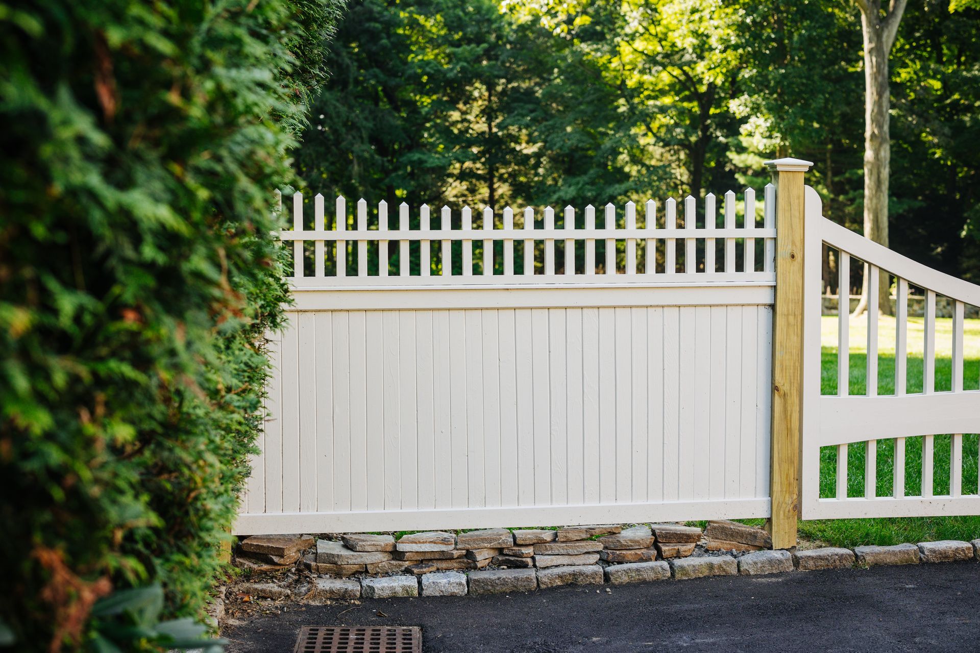 A white picket fence surrounds a driveway with trees in the background.