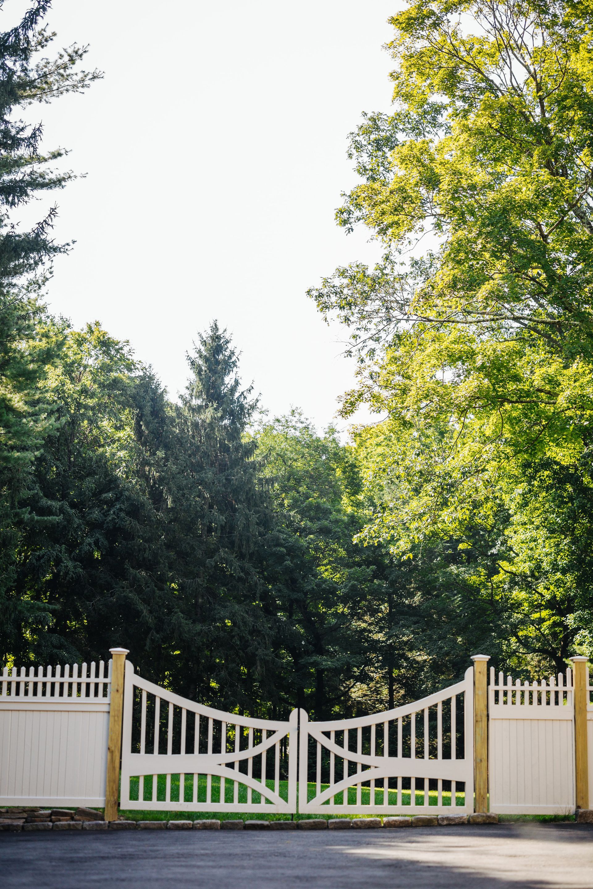 A white gate is open to a driveway surrounded by trees.