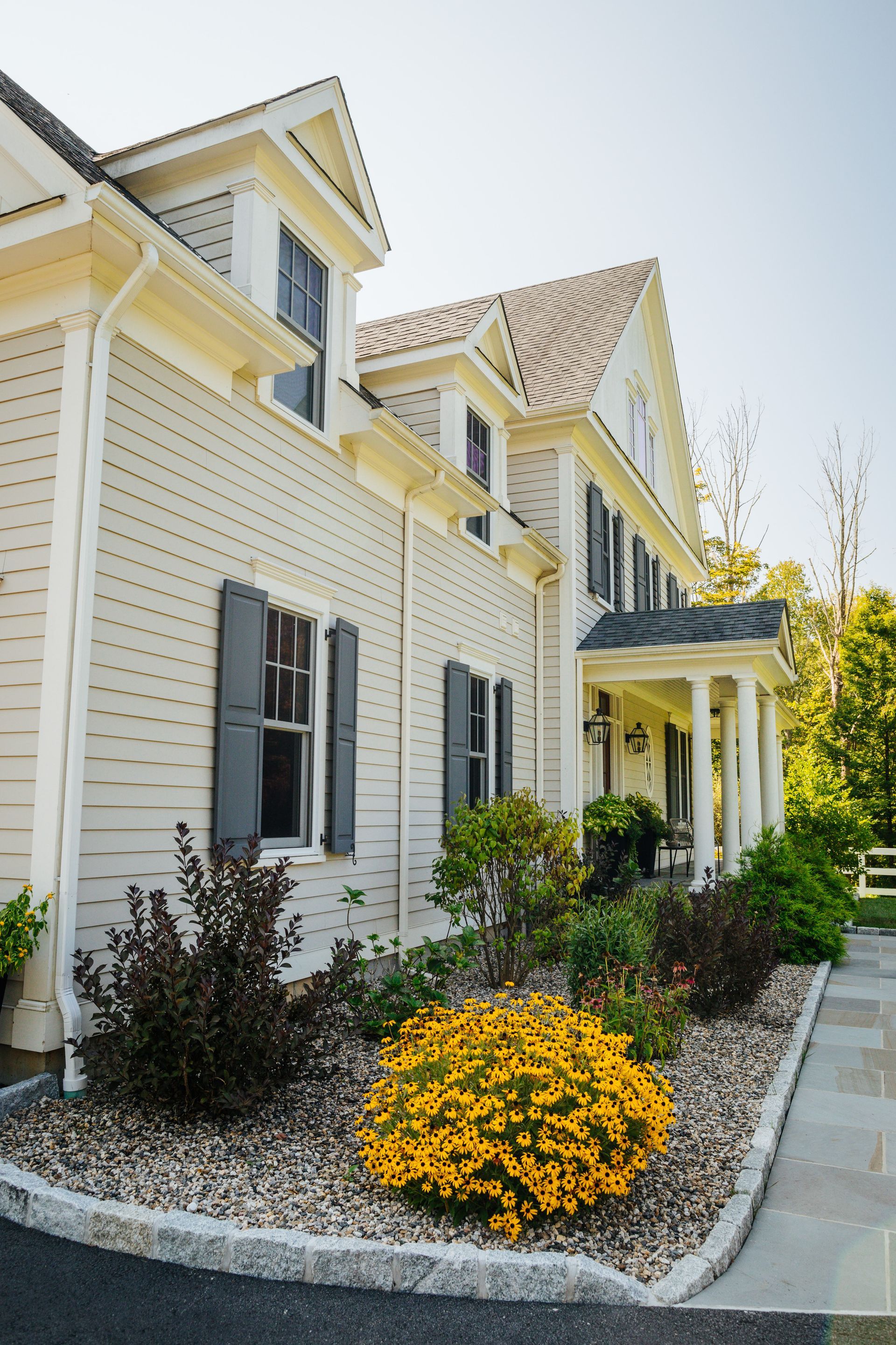 A large white house with gray shutters and yellow flowers in front of it.