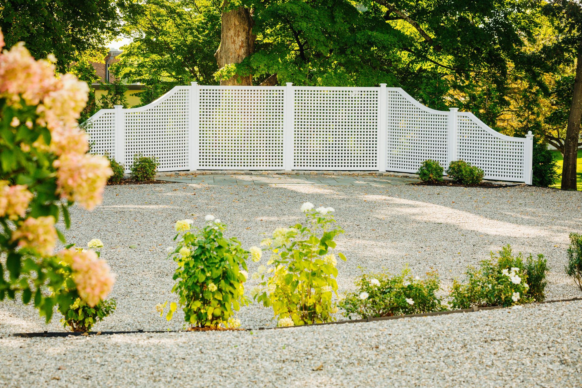 A gravel driveway with a white fence and flowers in the foreground.