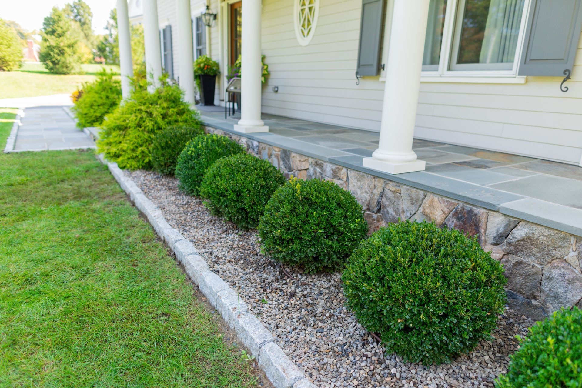 A white house with a stone porch and a row of bushes in front of it.