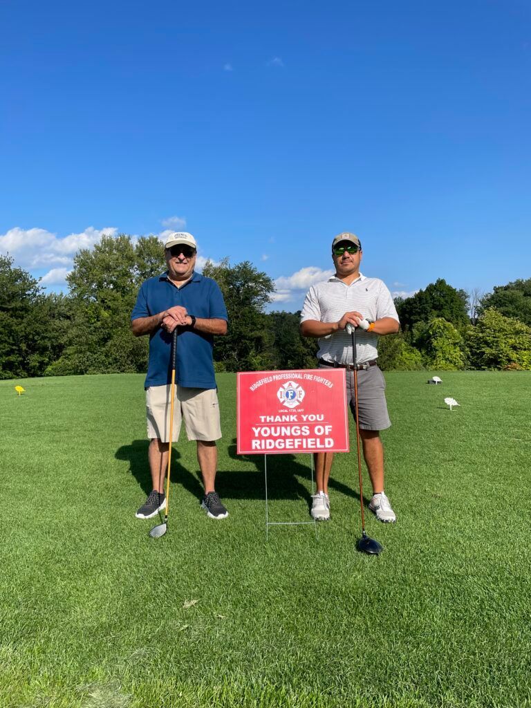 Two men are standing on a golf course holding golf clubs and a sign.