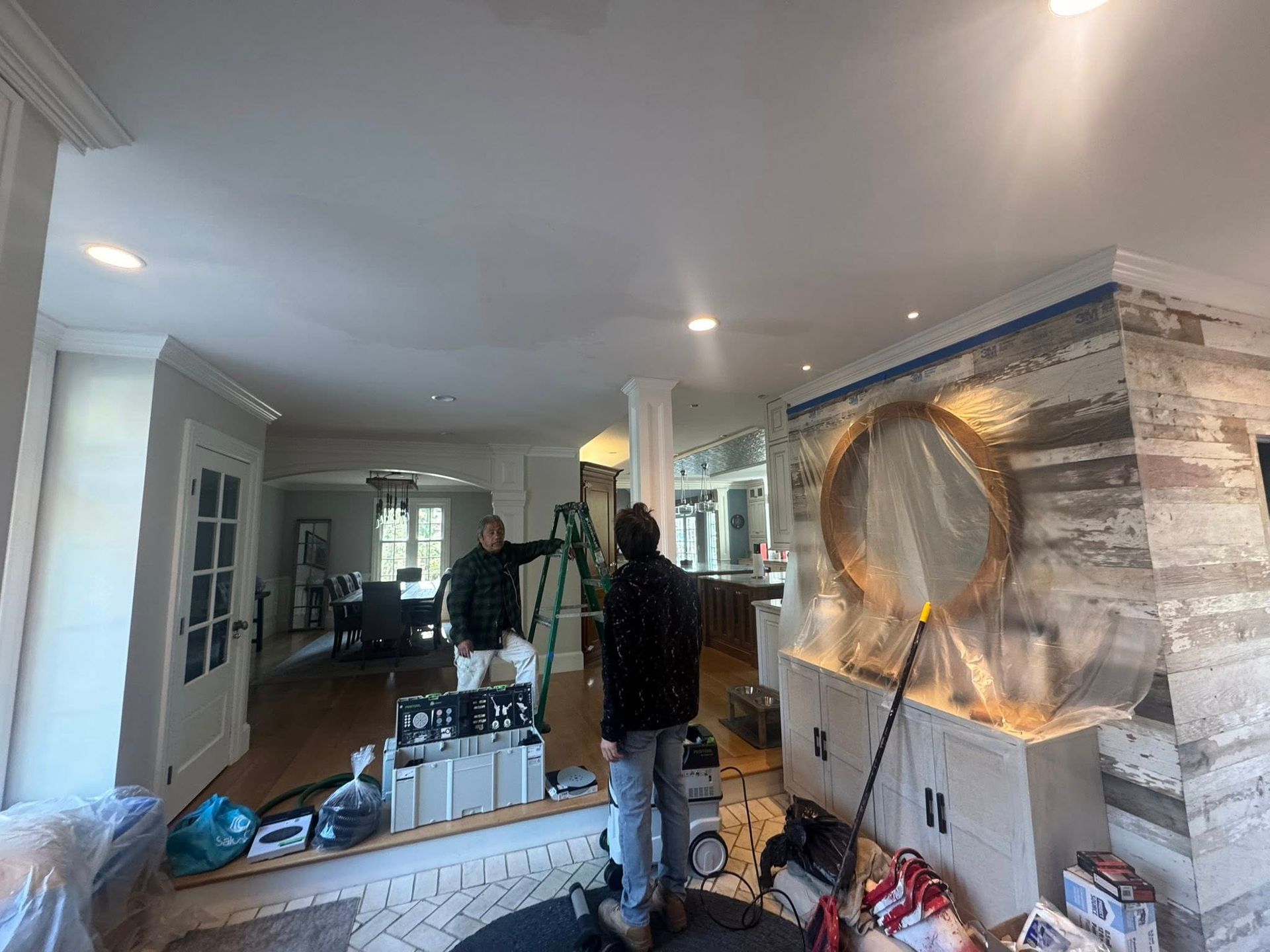 Hallway renovation with workers, tools, and a striped accent wall featuring a large round mirror.