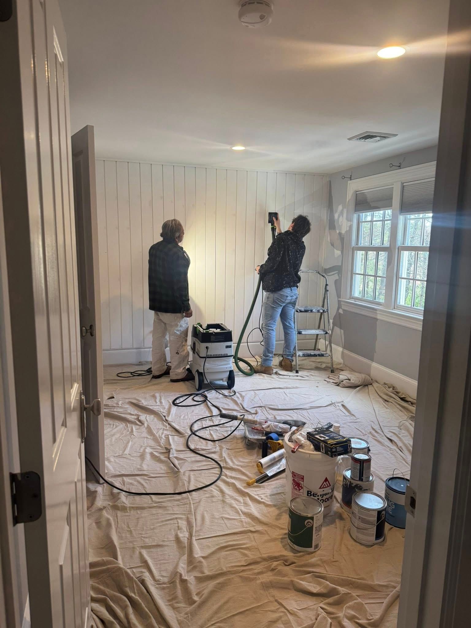 Two people painting a bright room with drop cloths, tools, and paint cans scattered on the floor.