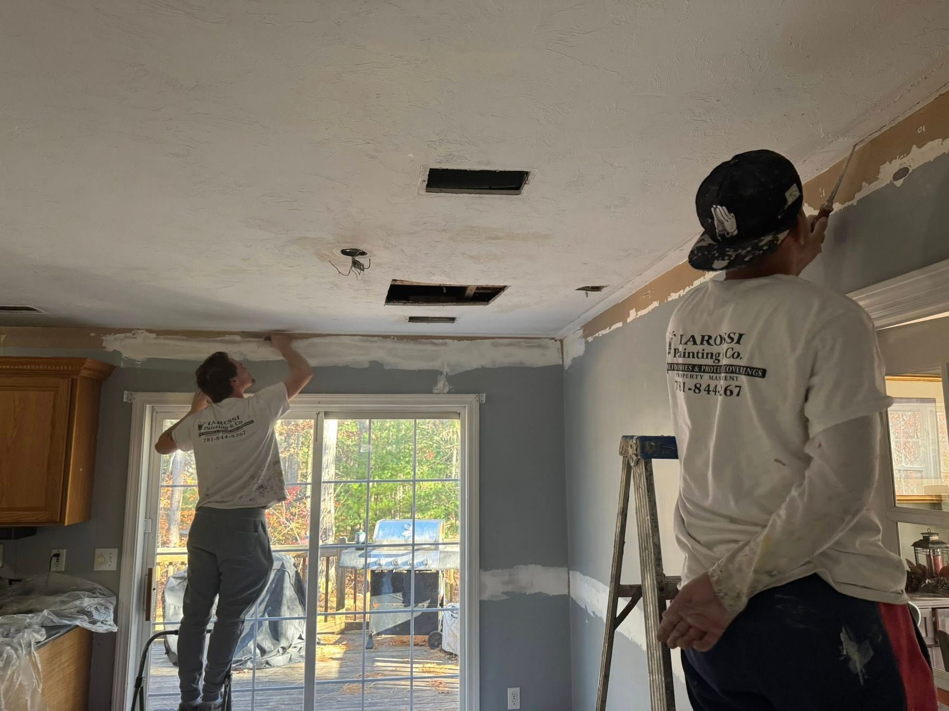 Two workers patching and painting a room ceiling near a window and ladder