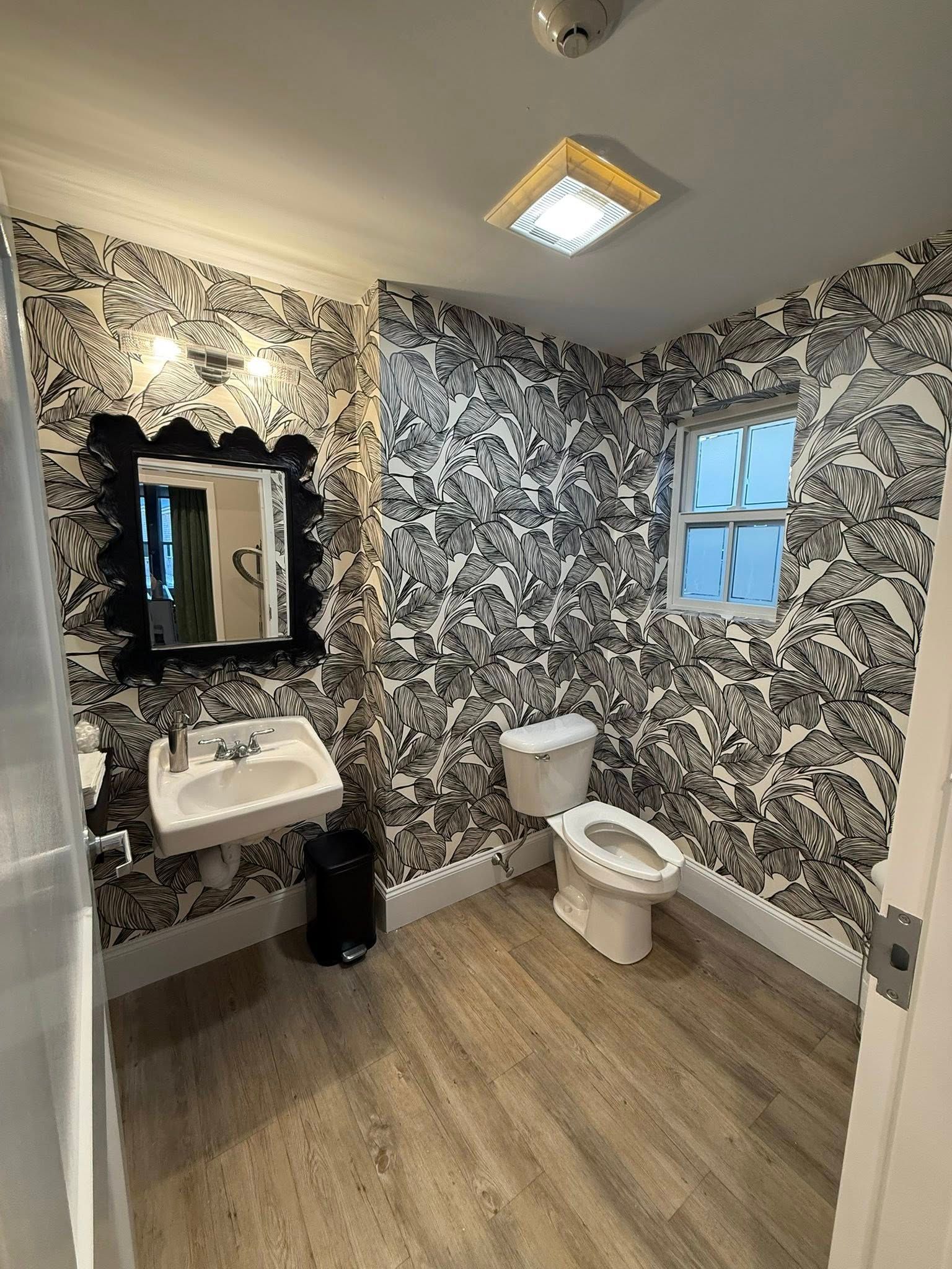 Small bathroom with patterned wallpaper, sink and mirror on left, toilet by window, and wood-look floor.