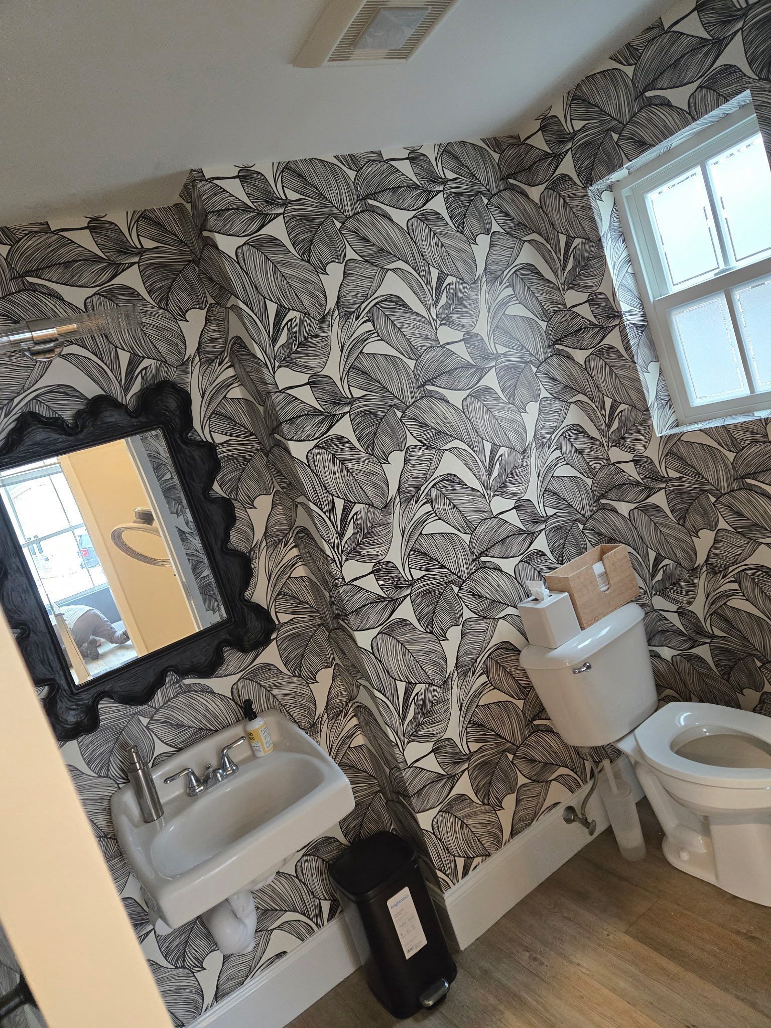 Small bathroom with black-and-white wallpaper, sink, toilet, mirror, and window.