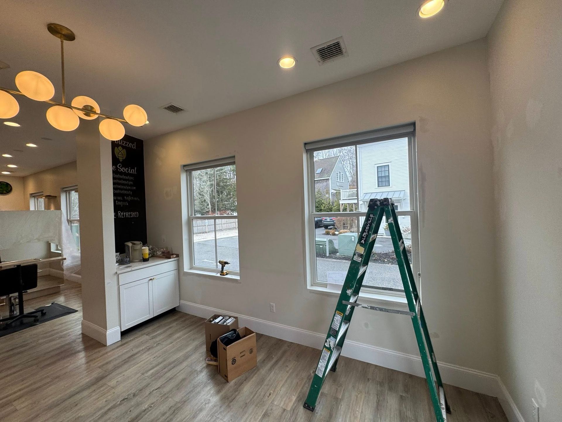 Bright living room with green ladder, white walls, large windows, and wood flooring under renovation