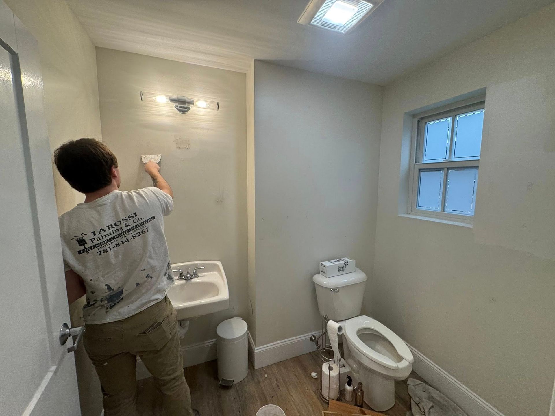 Worker painting a small white bathroom with a sink, toilet, and window.