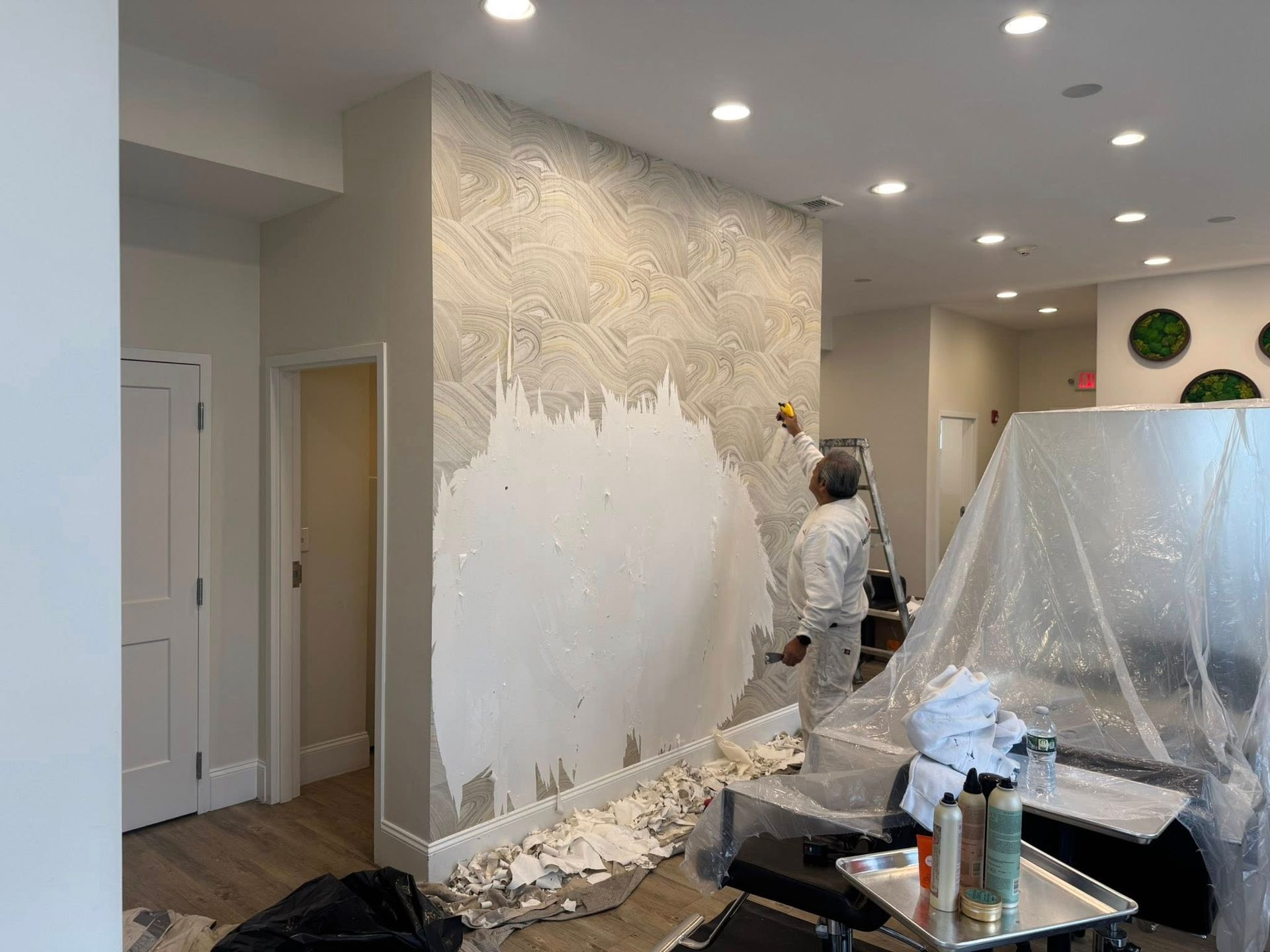 Person painting a wall in a bright room with plastic-covered furniture and paint supplies on the floor