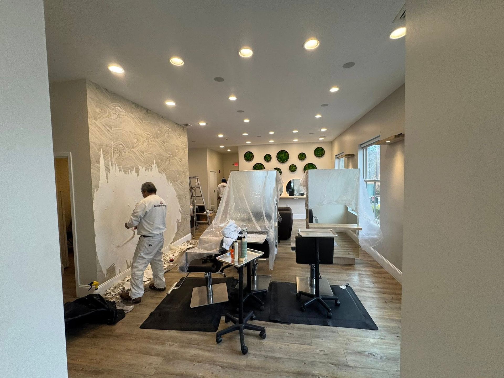 Two painters in a bright room with plastic-covered furniture, preparing walls for renovation.
