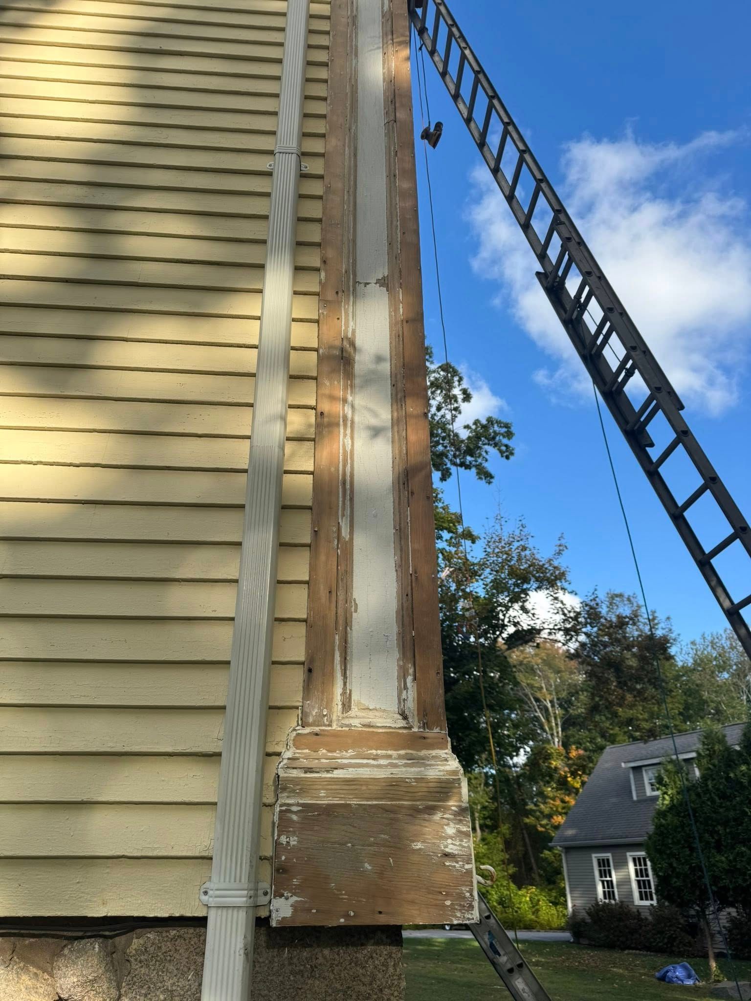 Damaged vertical wooden beam beside house siding, with a ladder leaning nearby under a blue sky.