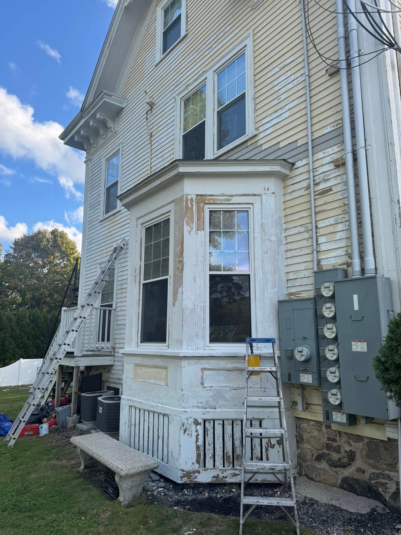 Side of a white house with utility meters, ladder, and peeling paint by a small porch