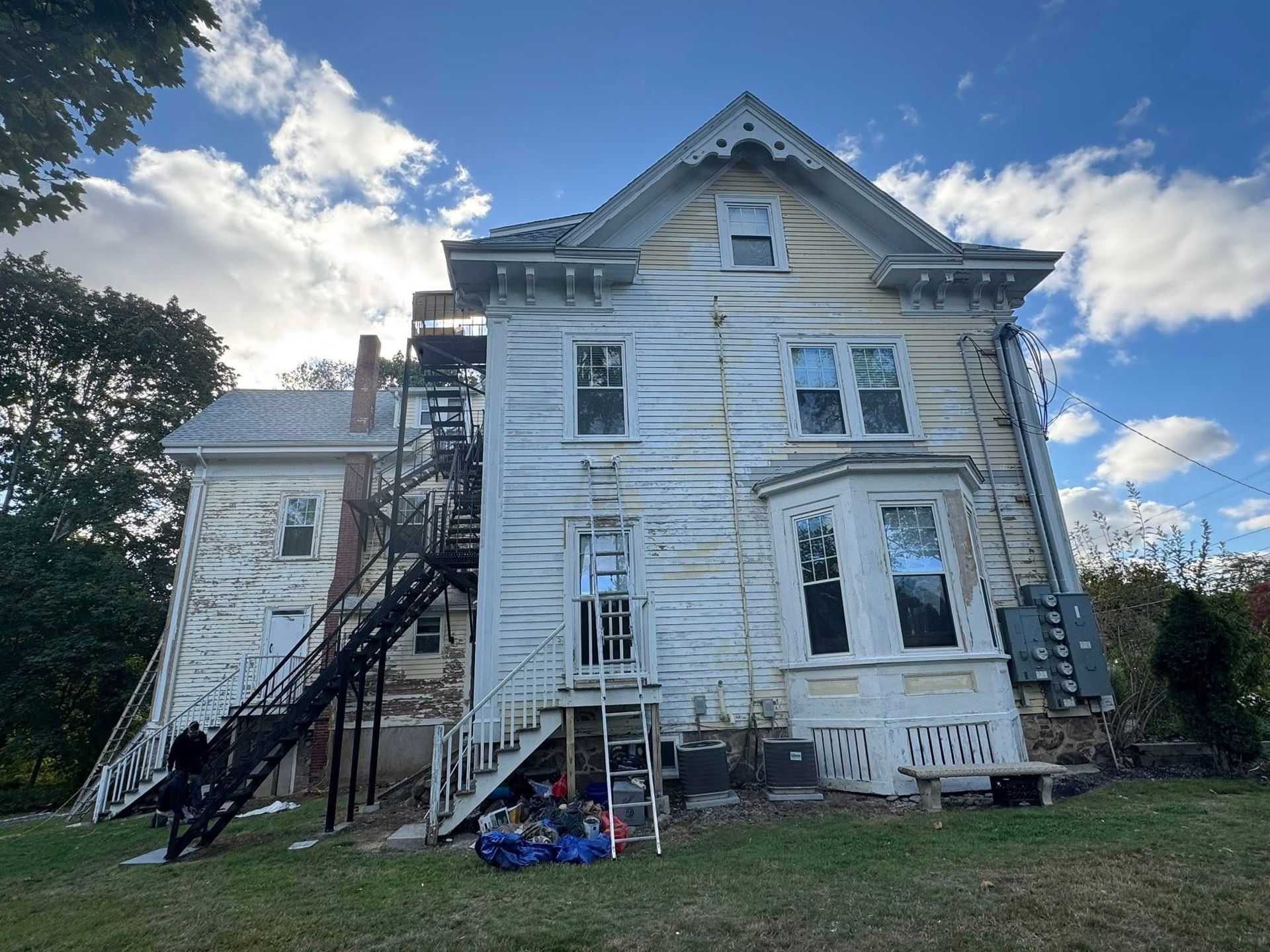 Old white house with peeling paint and exterior fire escape under a cloudy blue sky