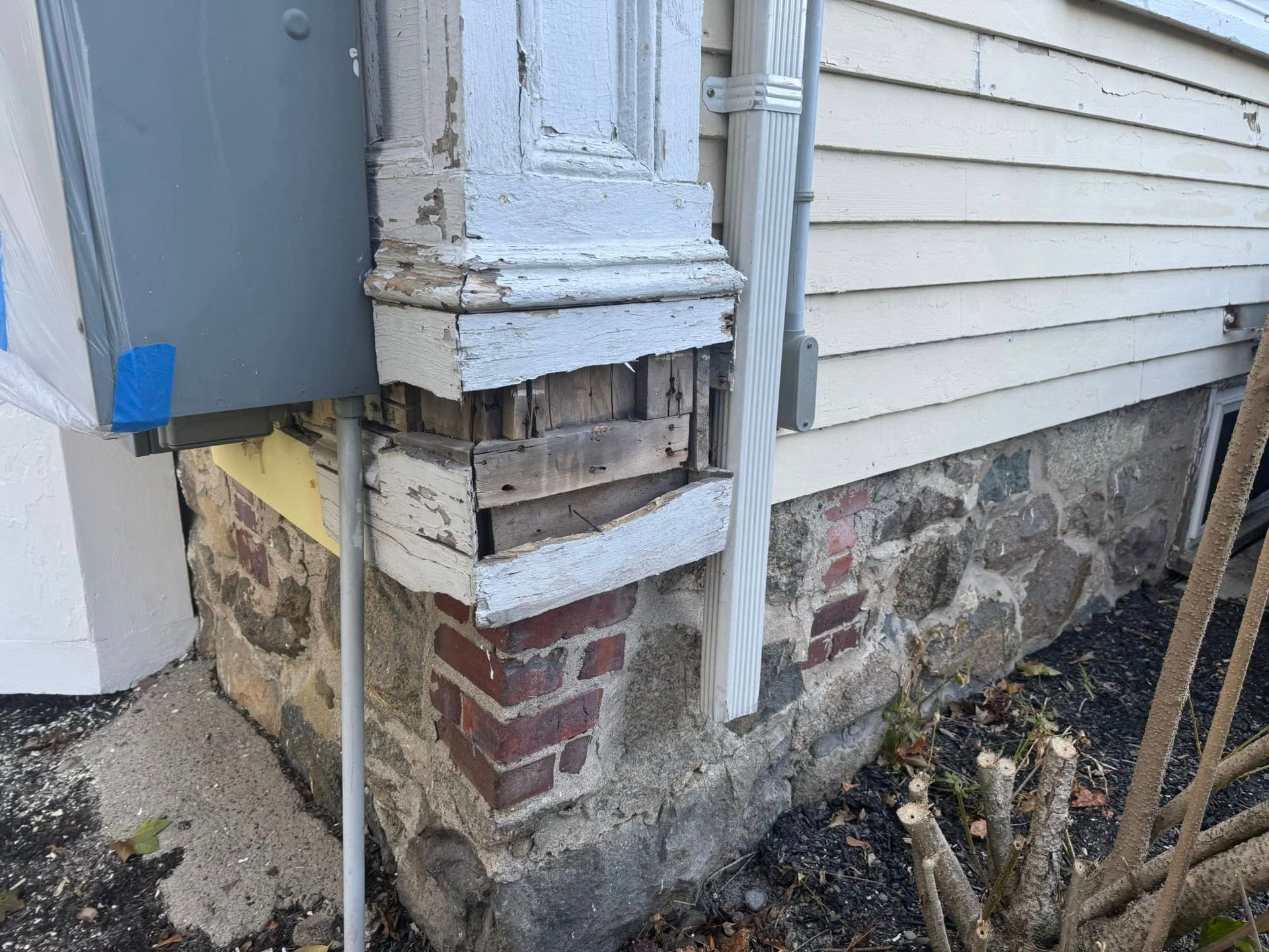 Damaged house corner with exposed framing, crumbling siding, and brick foundation near utility meter
